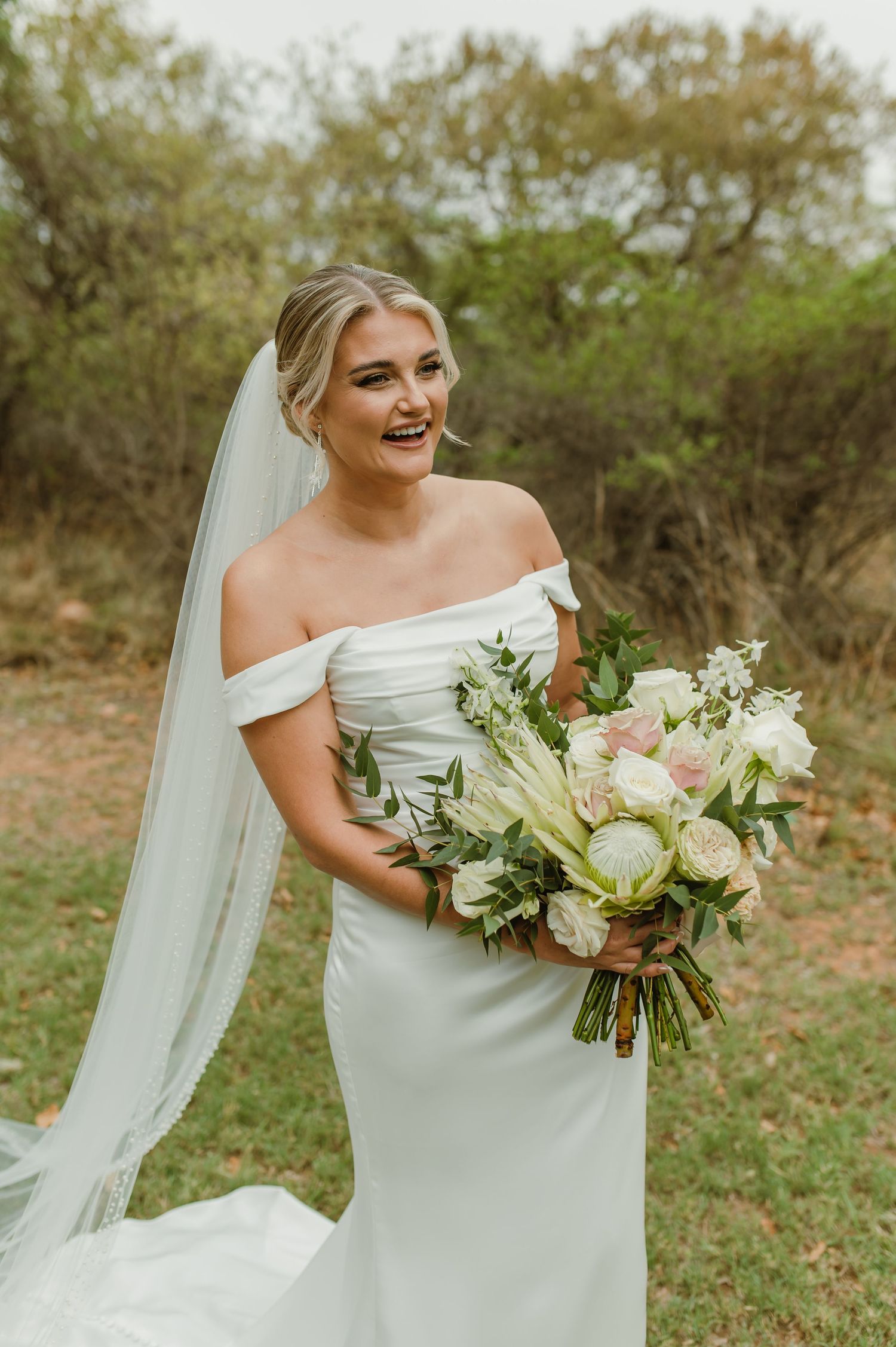 Bride in off-shoulder wedding dress smiles while holding a lush white and green bouquet outdoors.