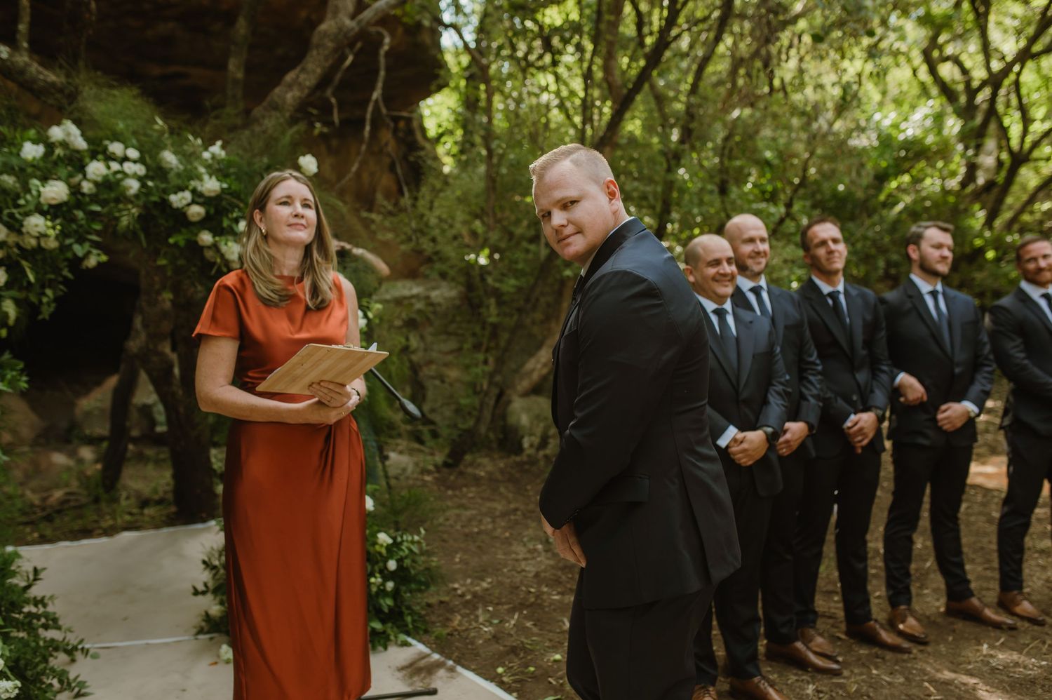 Groomsmen in dark suits stand in formation along a woodland ceremony site.