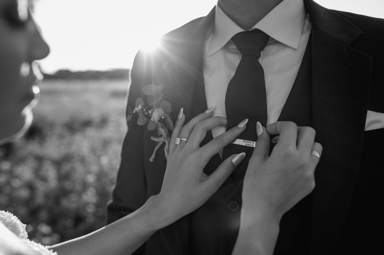 Close-up black and white detail of hands adjusting a groom's tie in natural light.