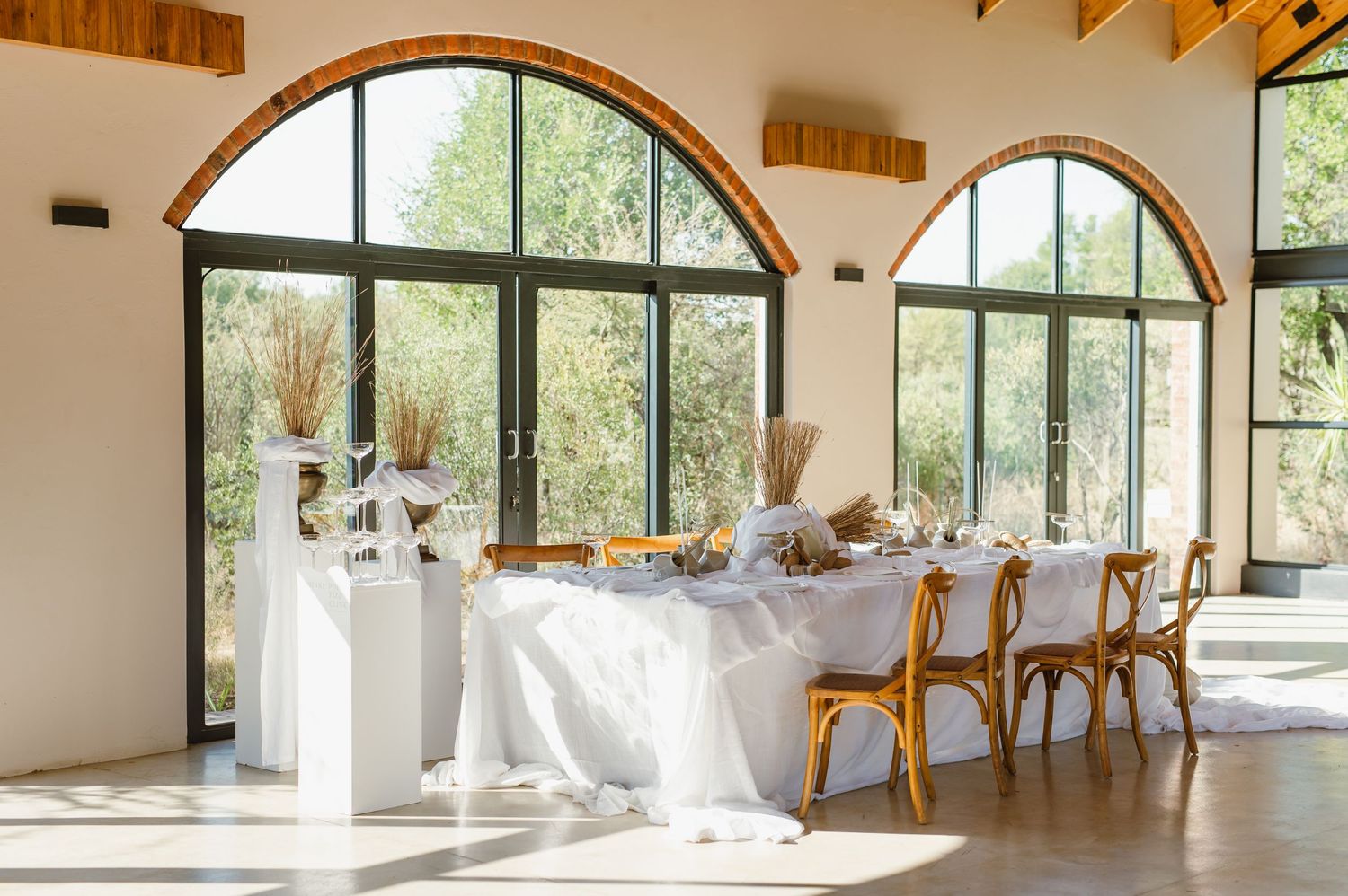 Modern wedding venue interior featuring wooden beams and large arched windows with natural lighting.