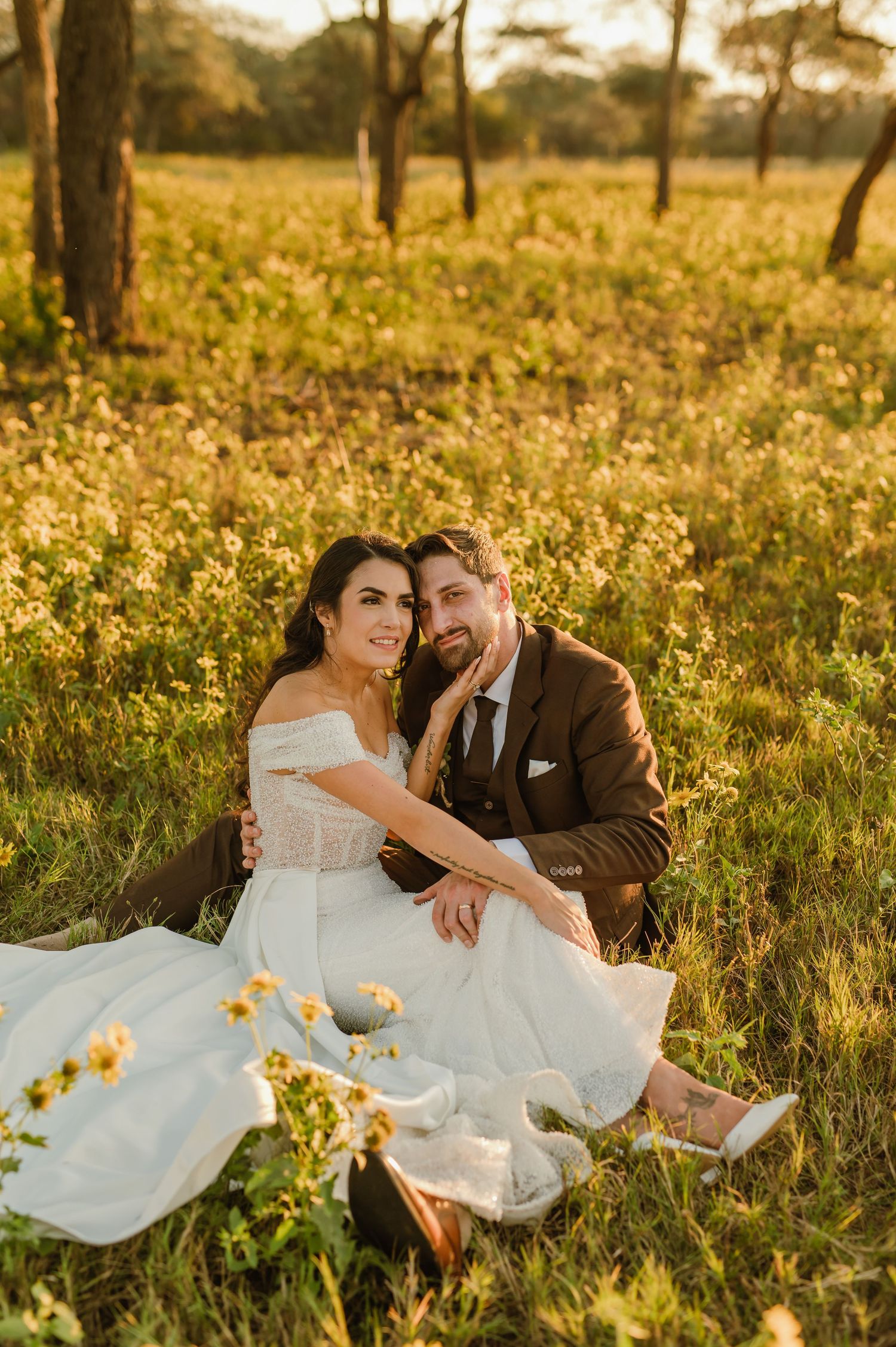 A couple shares a romantic moment in a golden wildflower field at sunset wearing wedding attire.