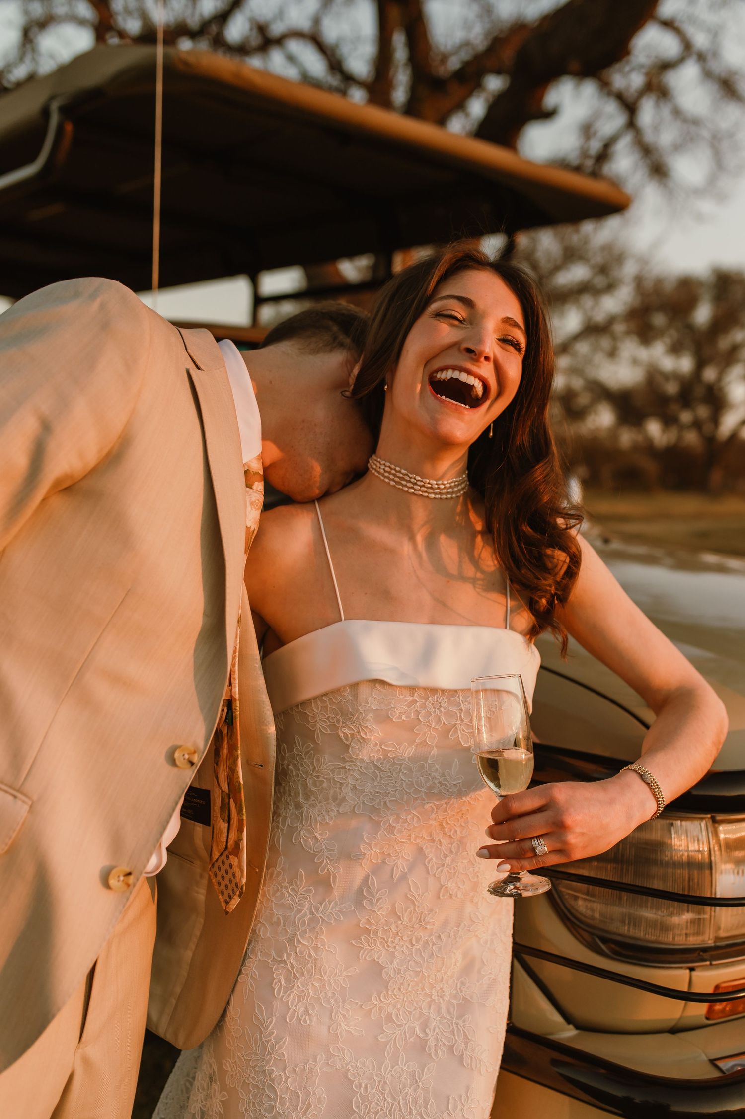 Laughing bride in white lace gown holds champagne while groom kisses her shoulder at sunset.