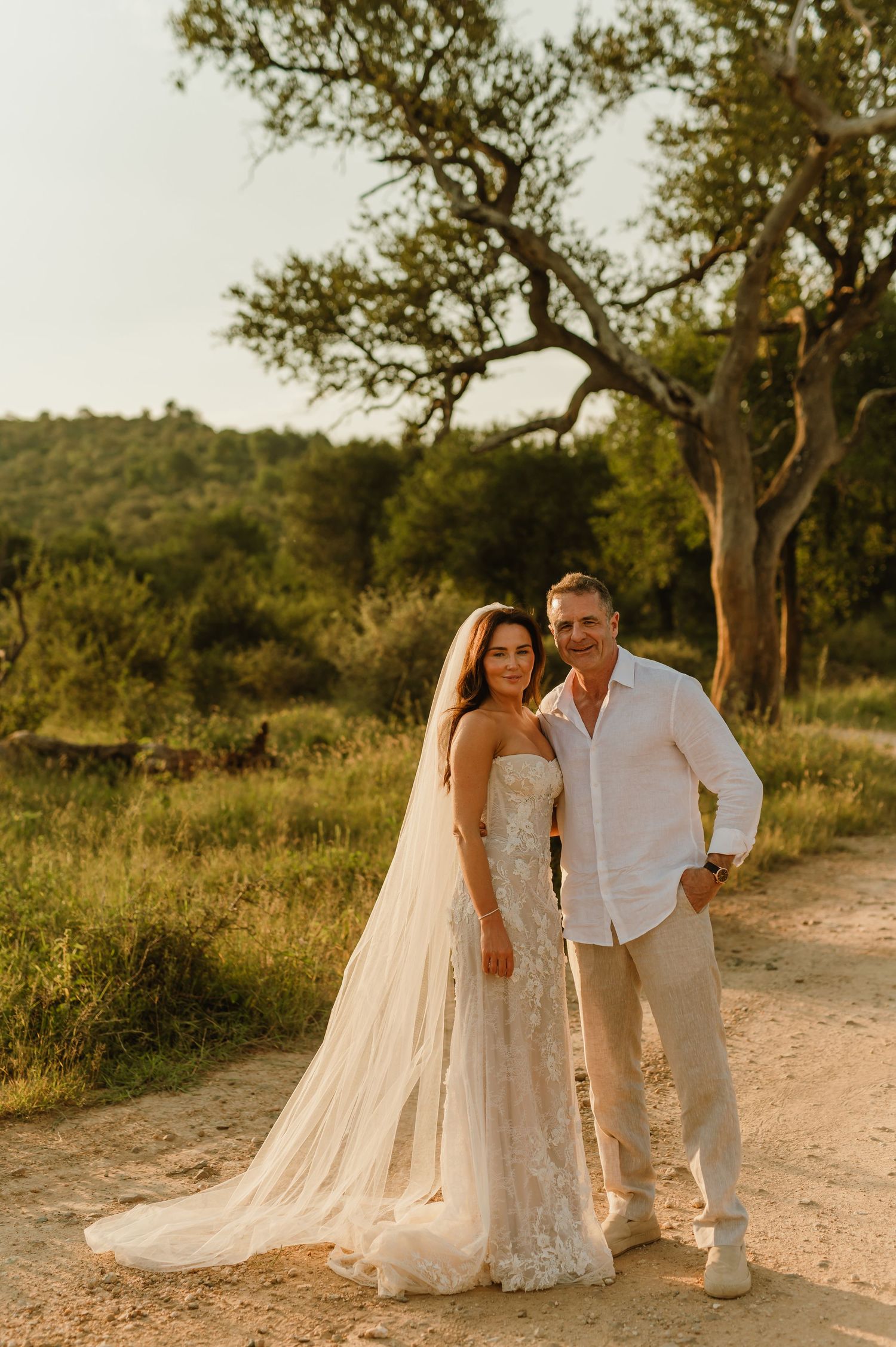 Bride in lace gown and groom in white shirt pose together on a dirt path surrounded by lush greenery at golden hour.