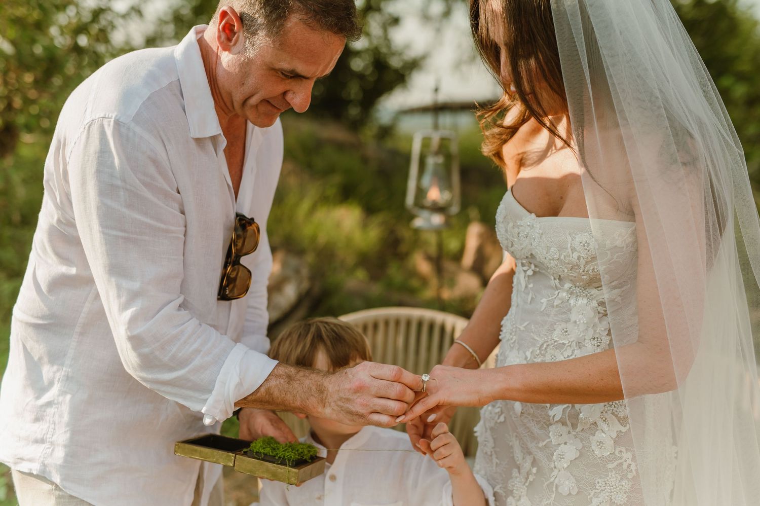 Groom placing wedding ring on bride's finger during outdoor ceremony, with child nearby, in elegant white attire.