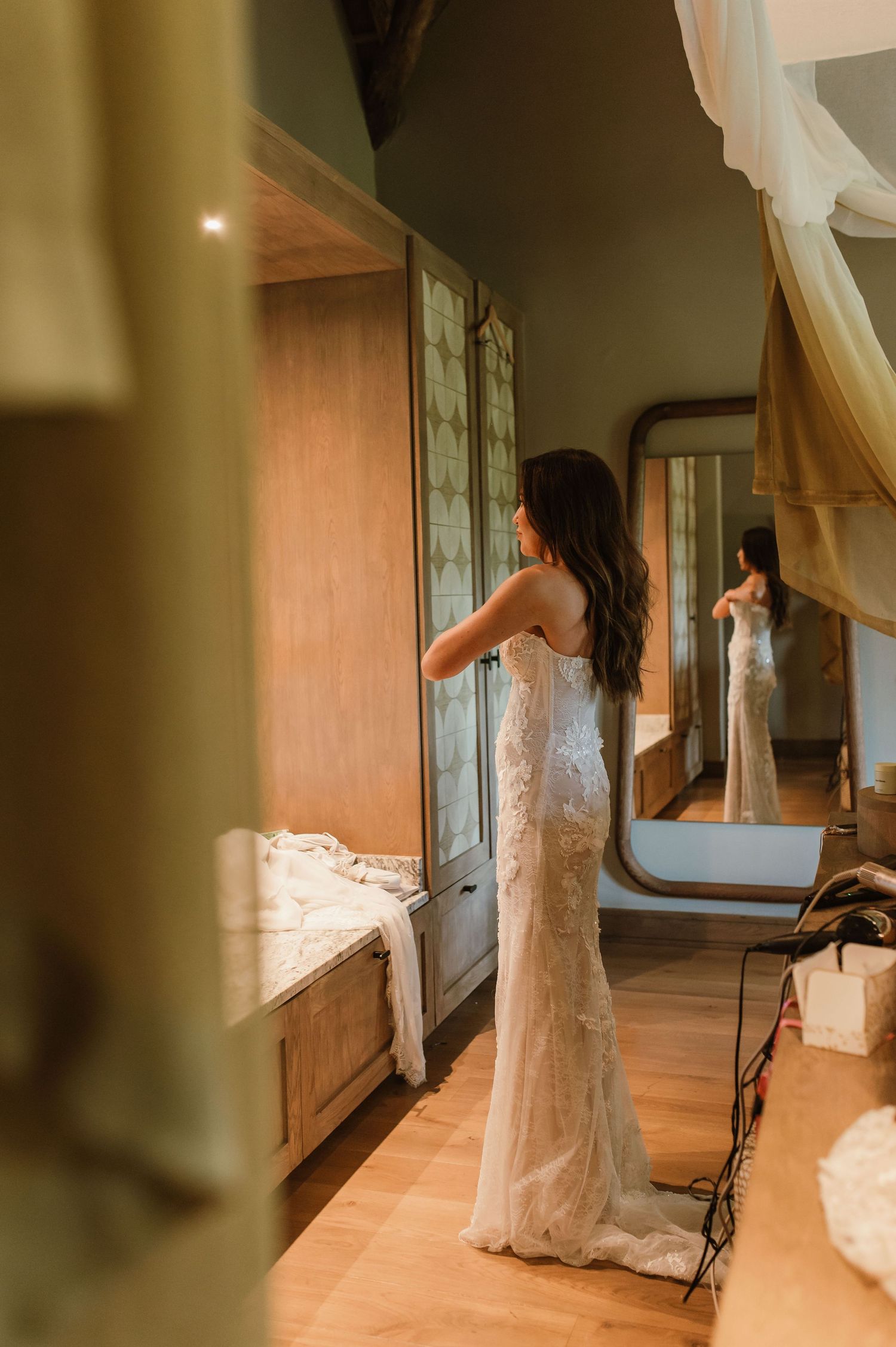 Bride in a fitted lace wedding dress admiring her reflection in a bedroom mirror.