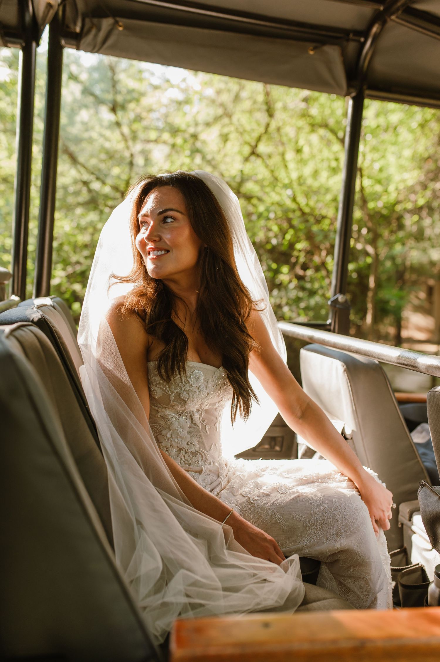 Smiling bride in strapless lace gown with veil seated inside vintage safari wedding vehicle.
