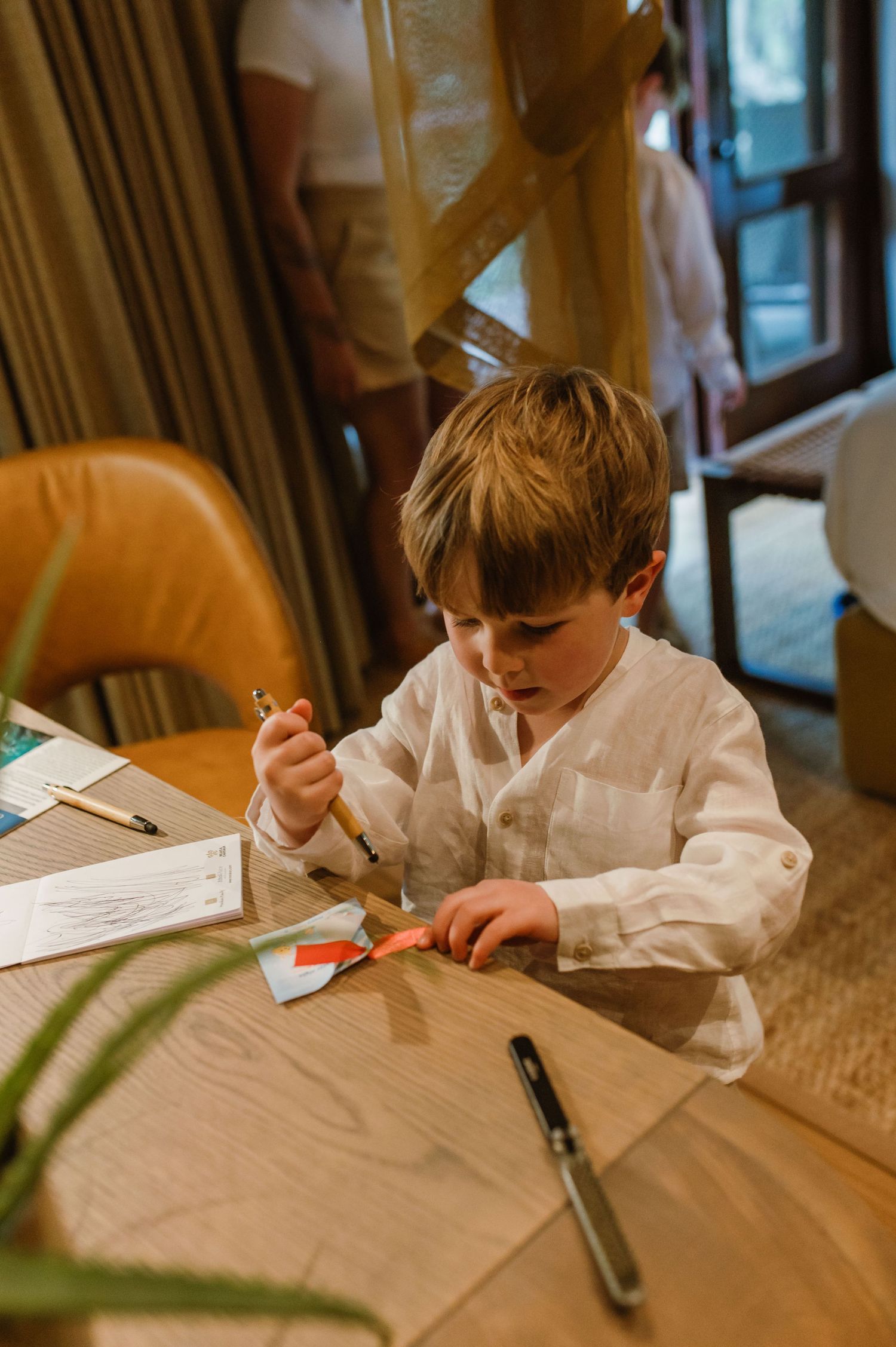 Young boy in white shirt crafting with scissors and paper at a wooden table.