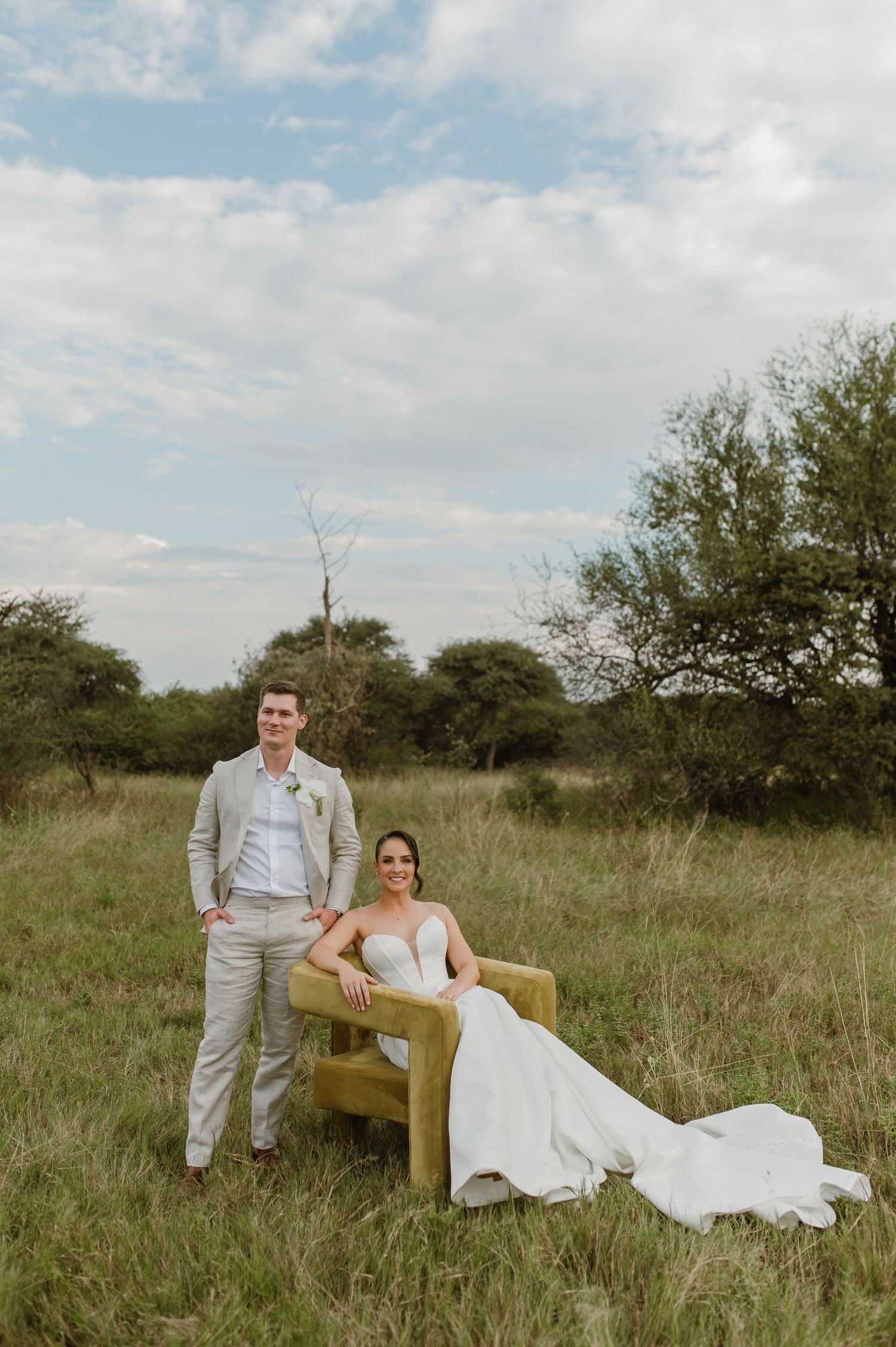 A newlywed couple poses with a vintage armchair in a rustic outdoor field setting.