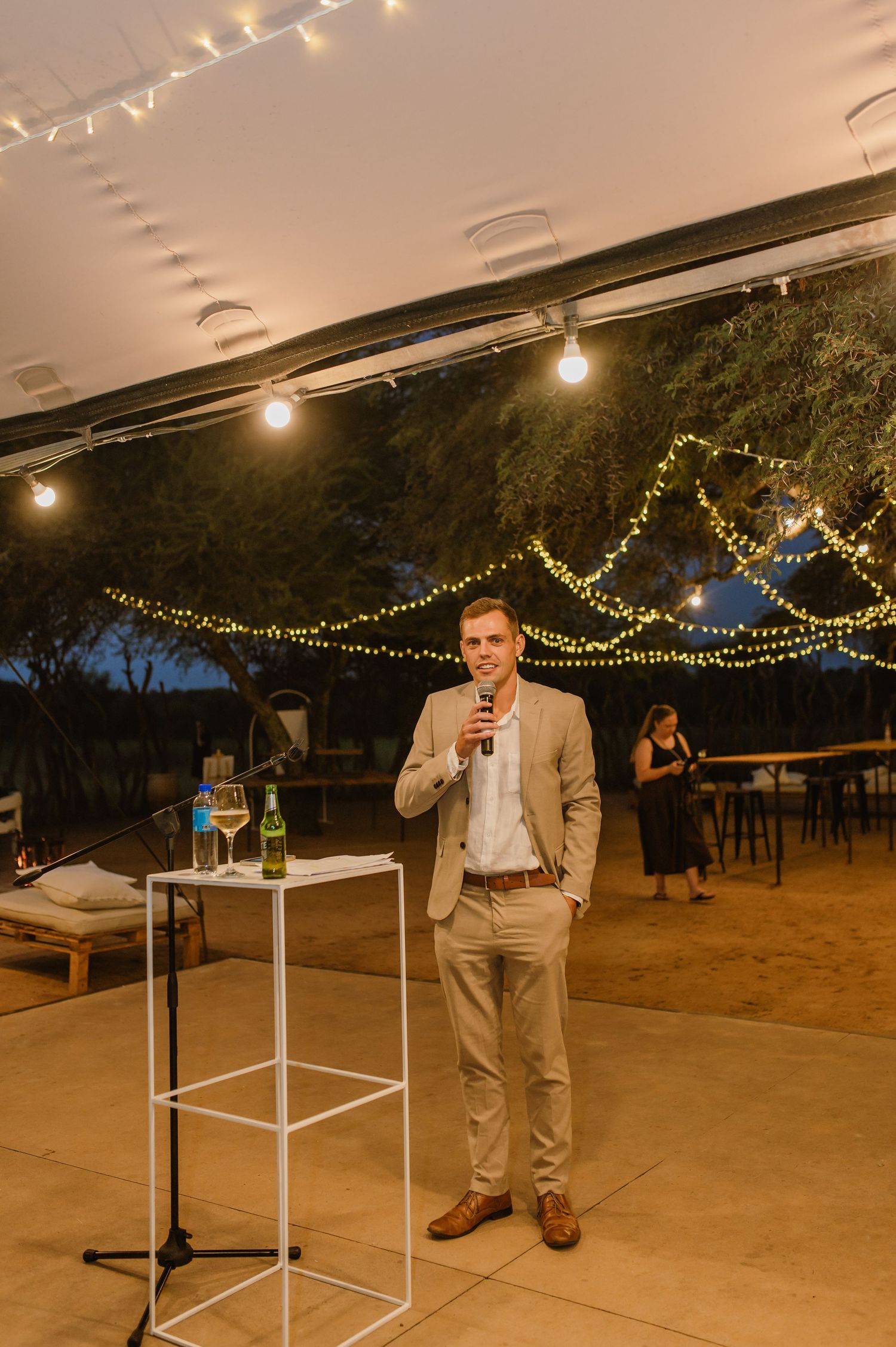 Speaker gives a toast at an outdoor evening reception under string lights and a covered patio area.