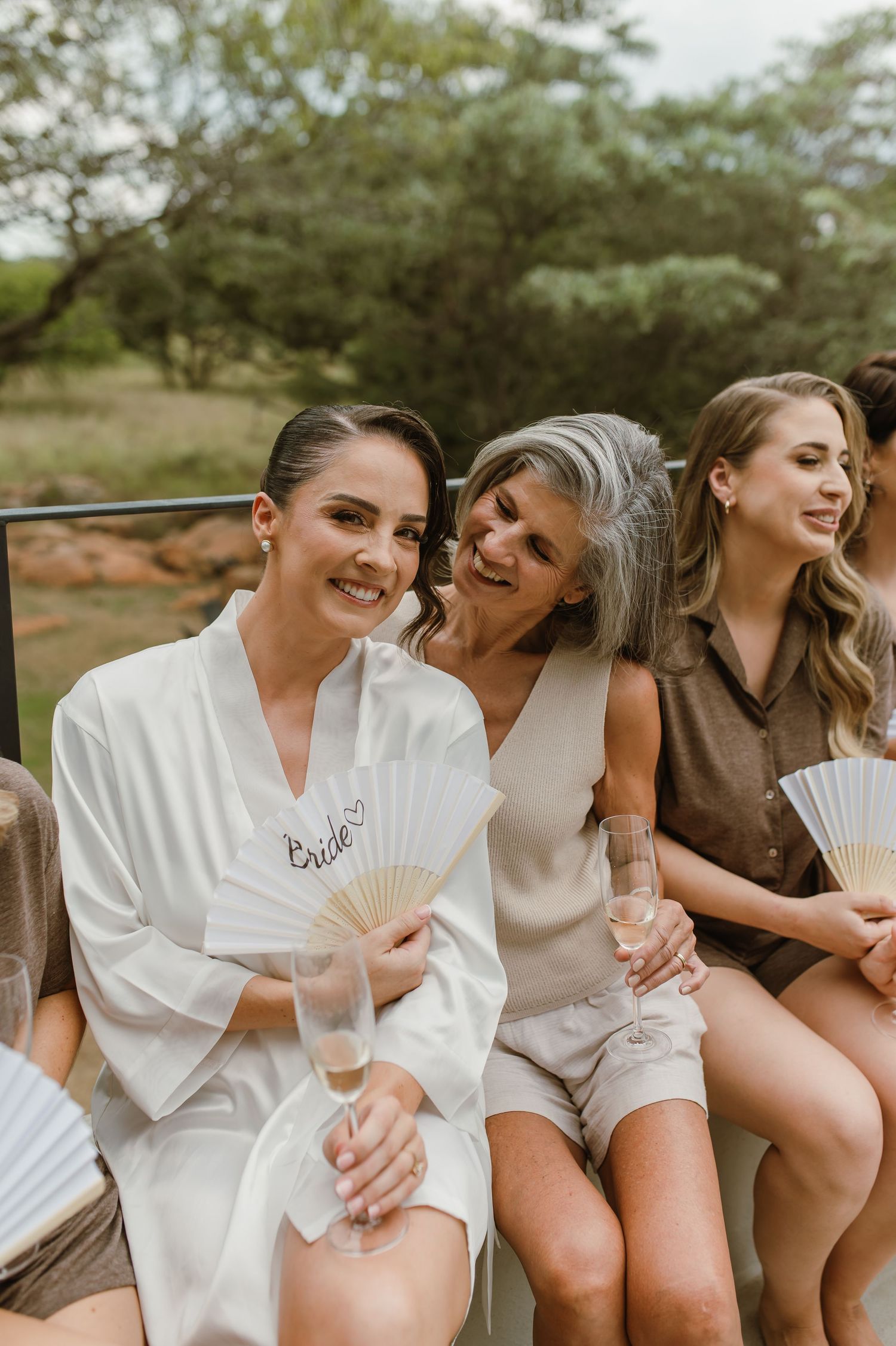 Wedding party members sharing laughs while holding paper fans outdoors.