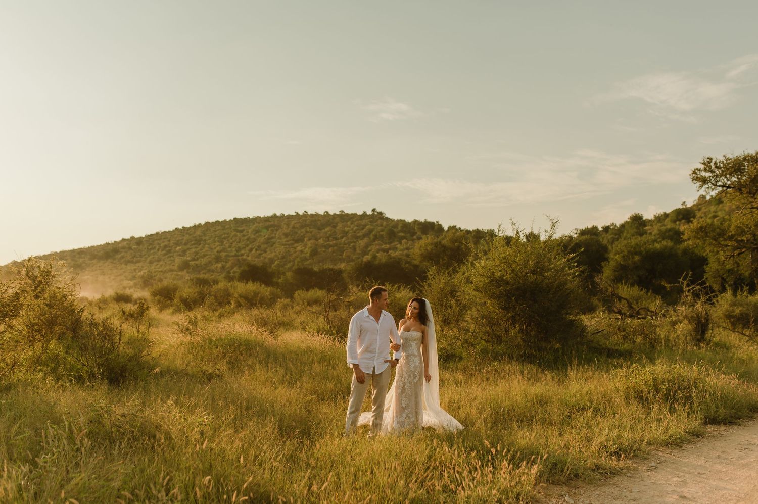 Bride and groom standing together in a golden sunlit meadow with rolling green hills in the background.