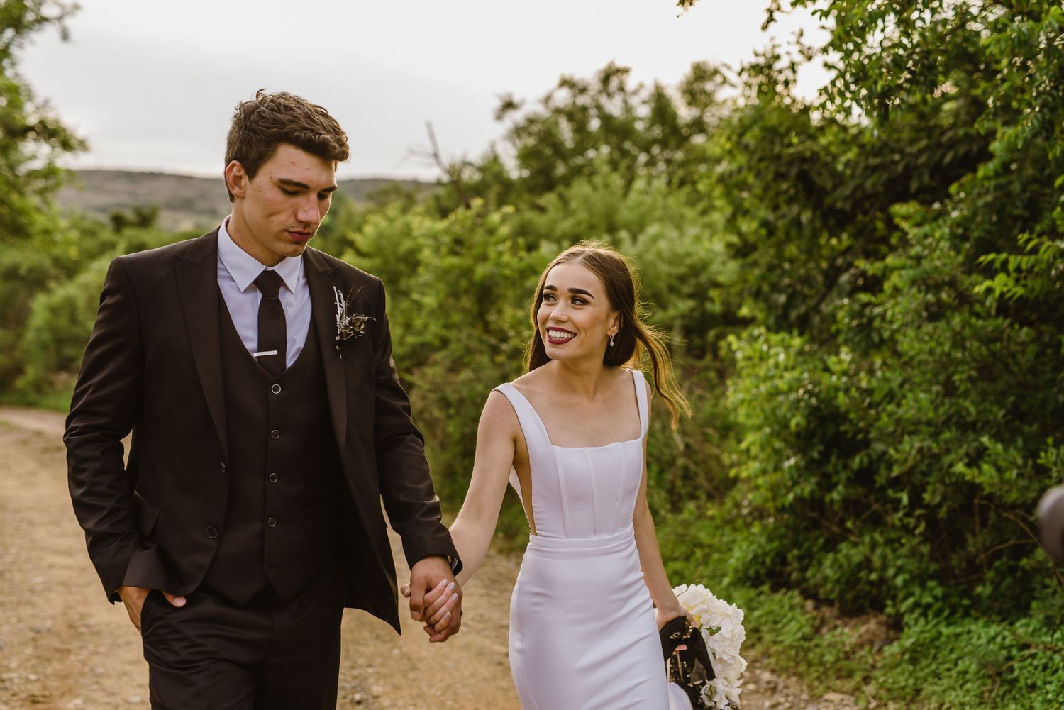 Wedding couple walks together on dirt path surrounded by green foliage.
