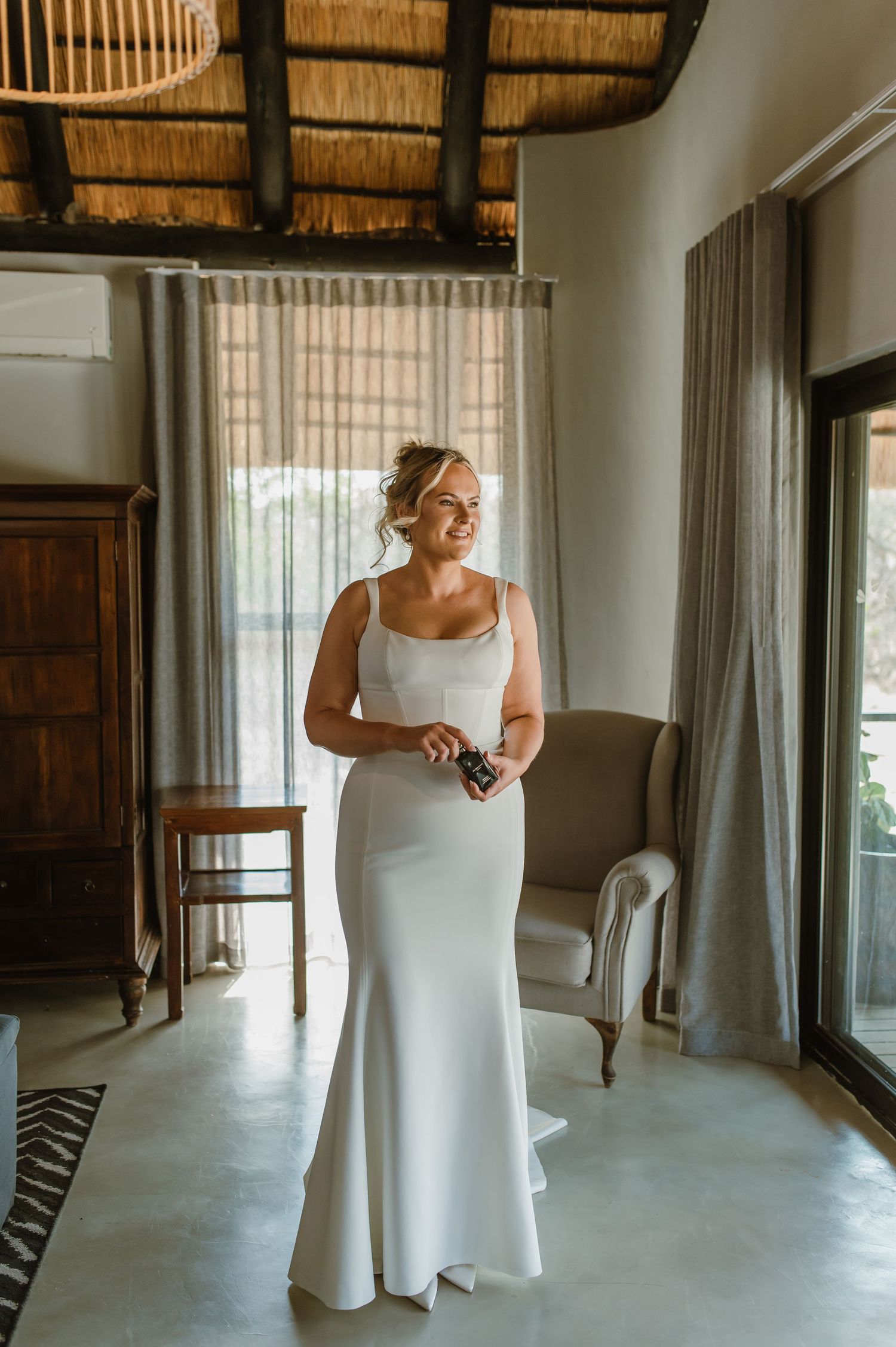 A person in a white wedding dress stands in a room with thatched roof and wooden furniture.