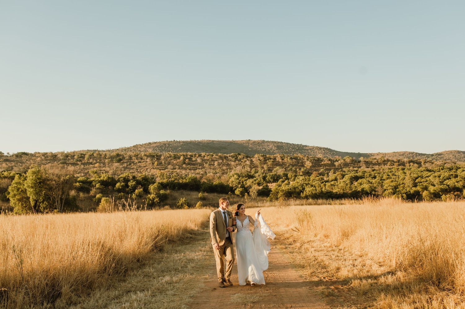 Wedding couple walks hand in hand through scenic field with rolling hills in background.