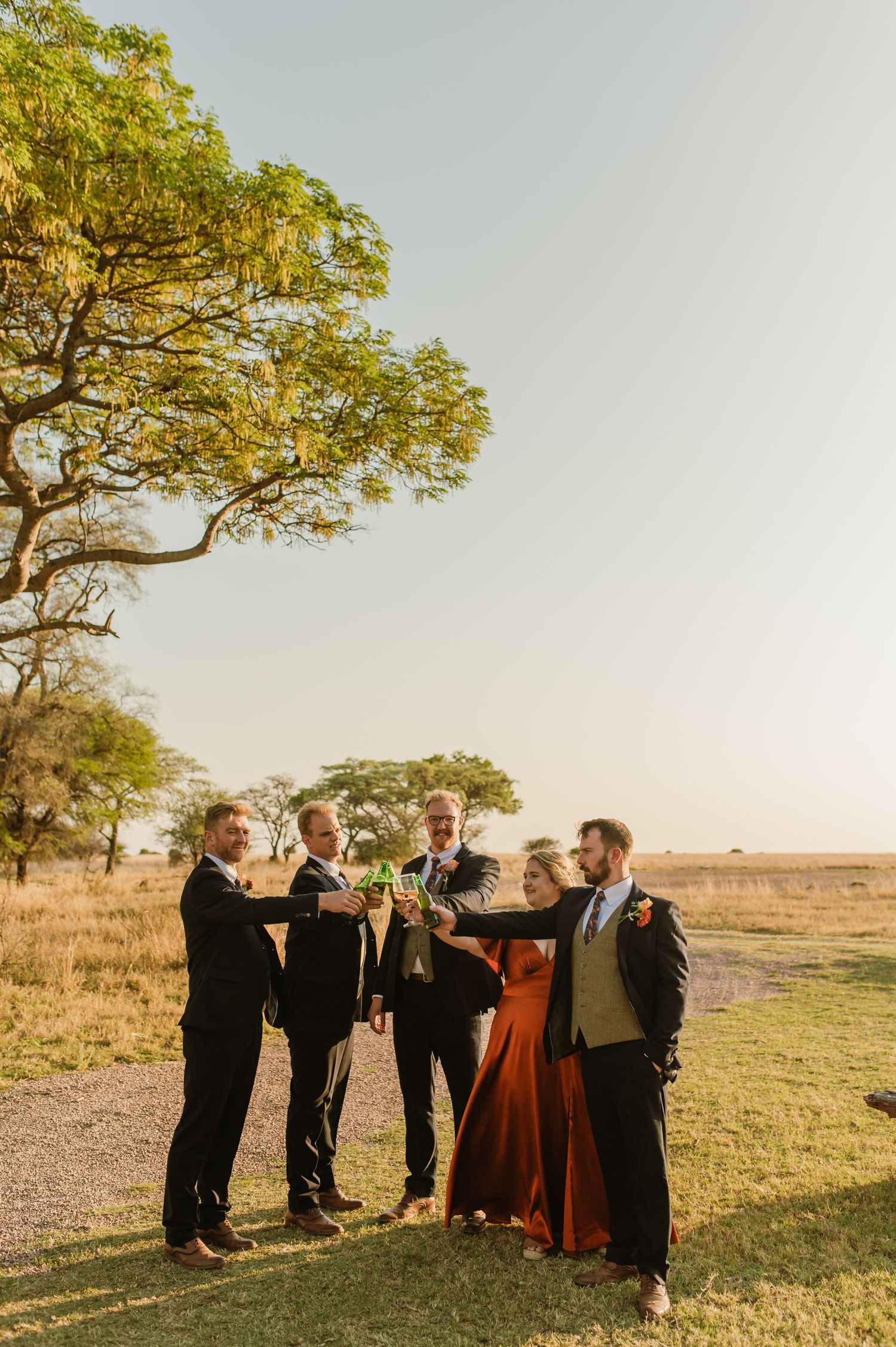 Wedding guests and party members share drinks under a tree in an outdoor setting.