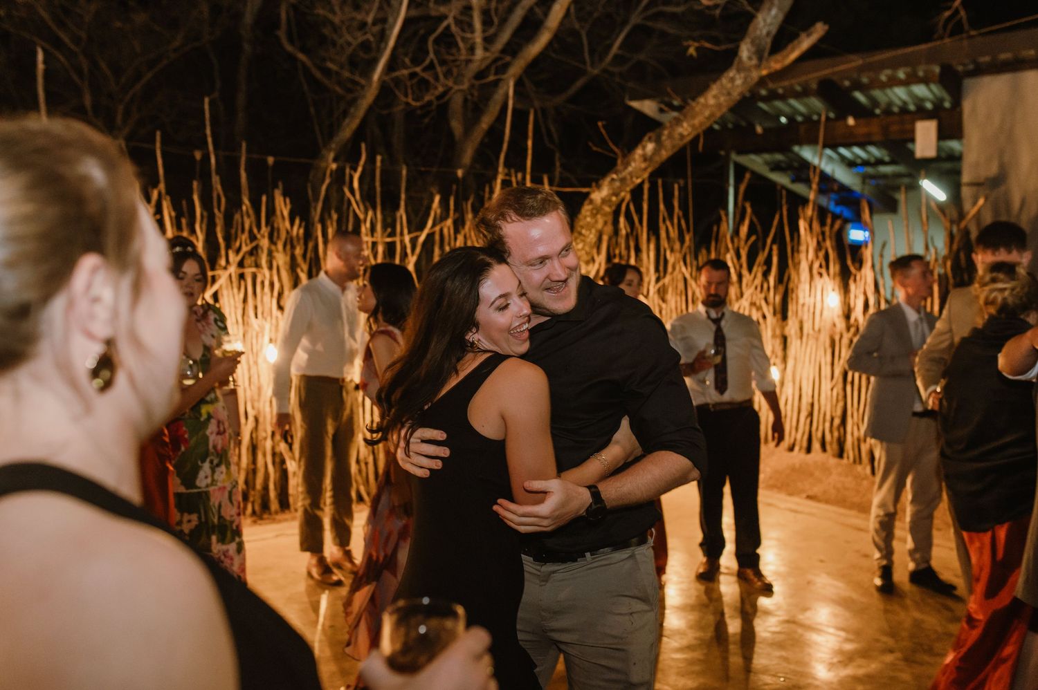 Wedding party dancing together in a rustic venue with thatched roof and ambient lighting.