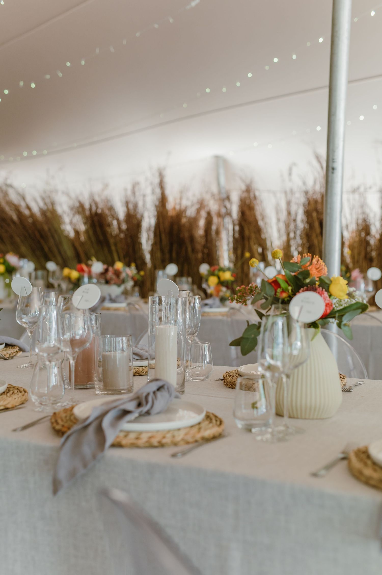 Long banquet table with string lights overhead and rustic dried grass backdrop creating an intimate atmosphere.