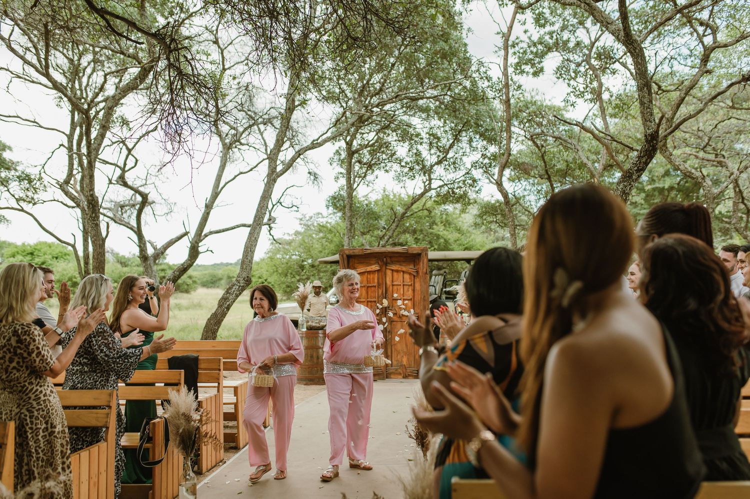 Wedding guests seated on wooden benches at an outdoor ceremony with trees in the background.
