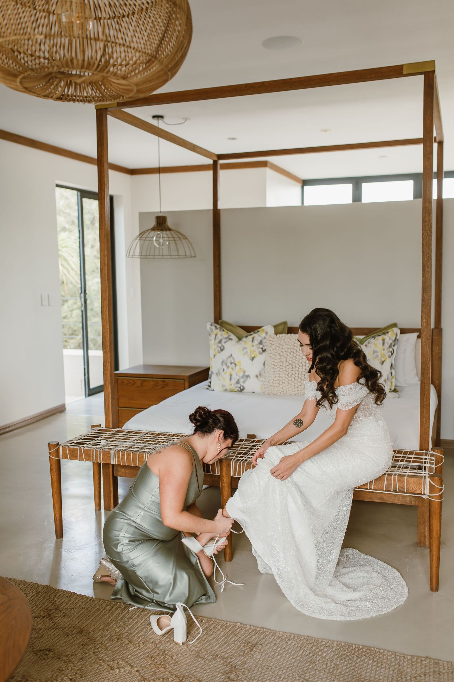 People adjusting a white dress near a wooden canopy bed in a bright, modern bedroom.