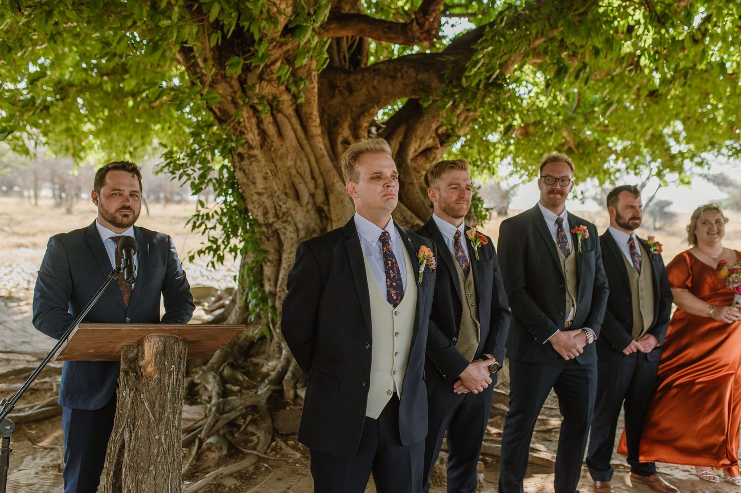 Wedding party members stand in formal attire under green leafy trees during an outdoor ceremony.