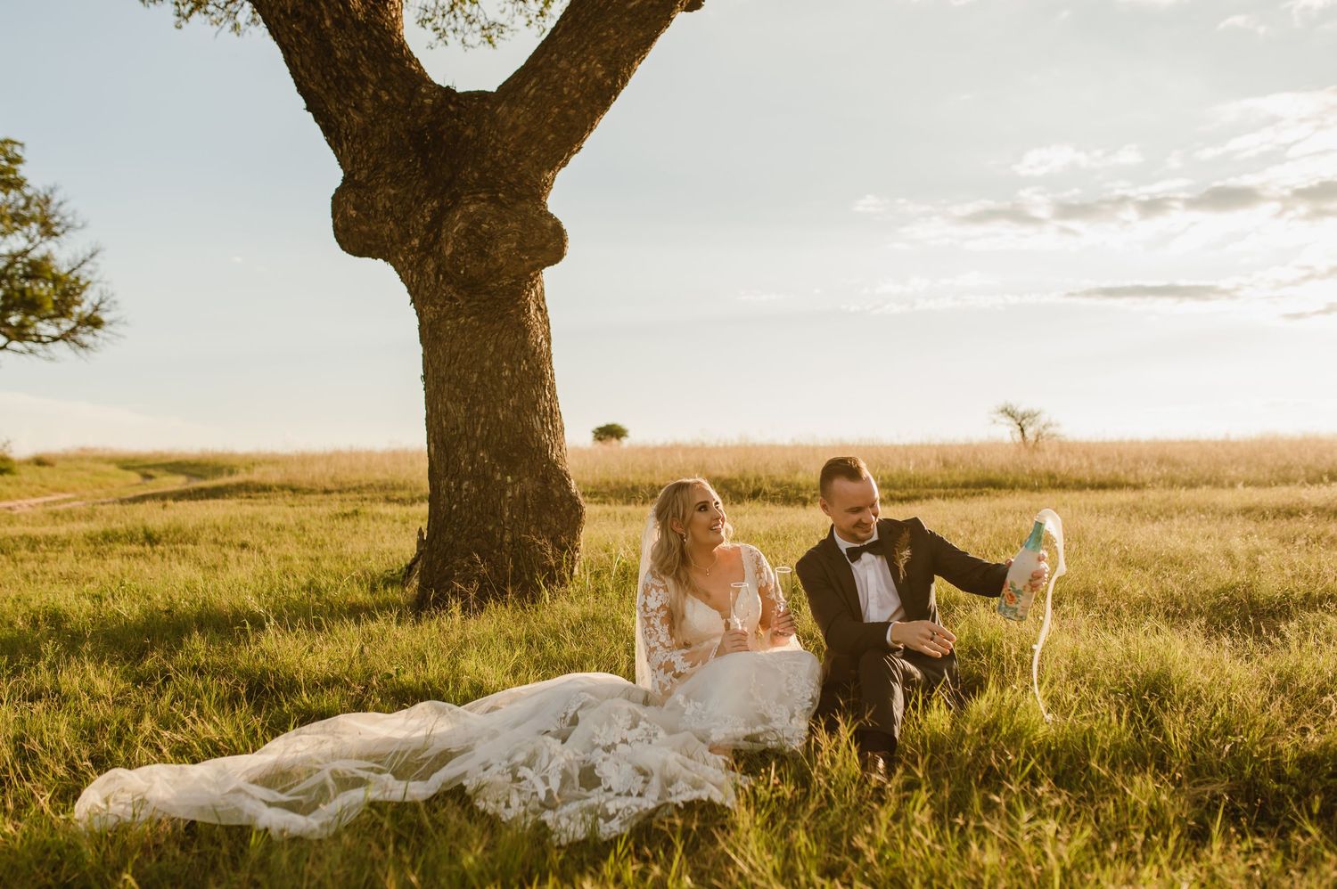 Newlyweds celebrate joyfully under a large tree in an open field during golden hour.