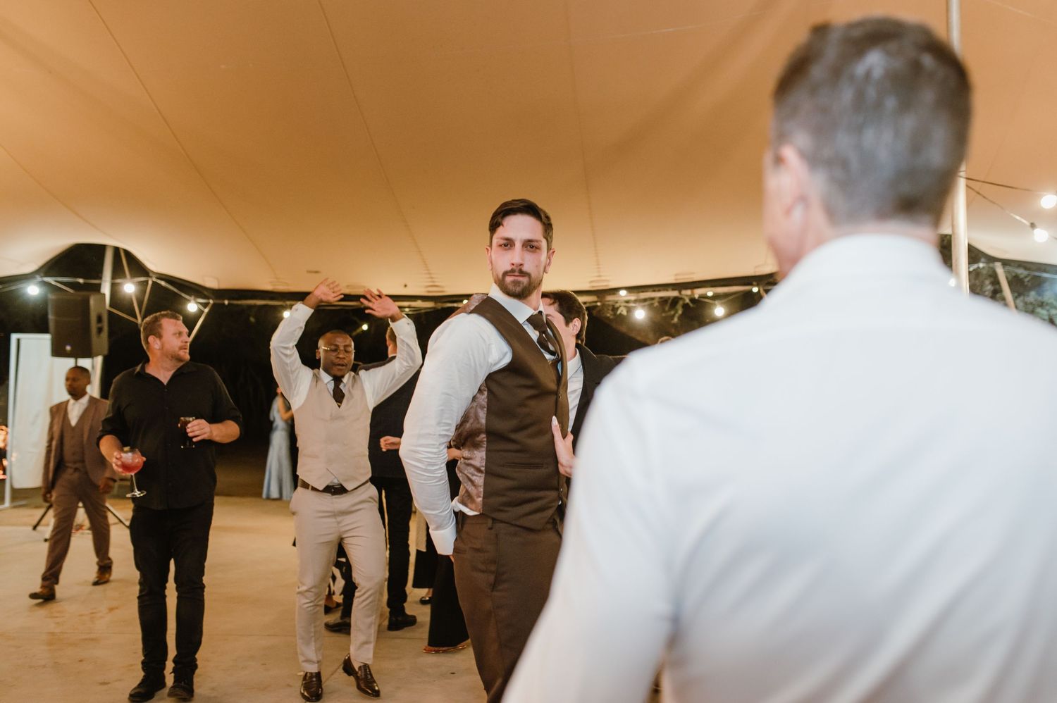 A person in a vest stands confidently on the dance floor during a tented wedding reception.