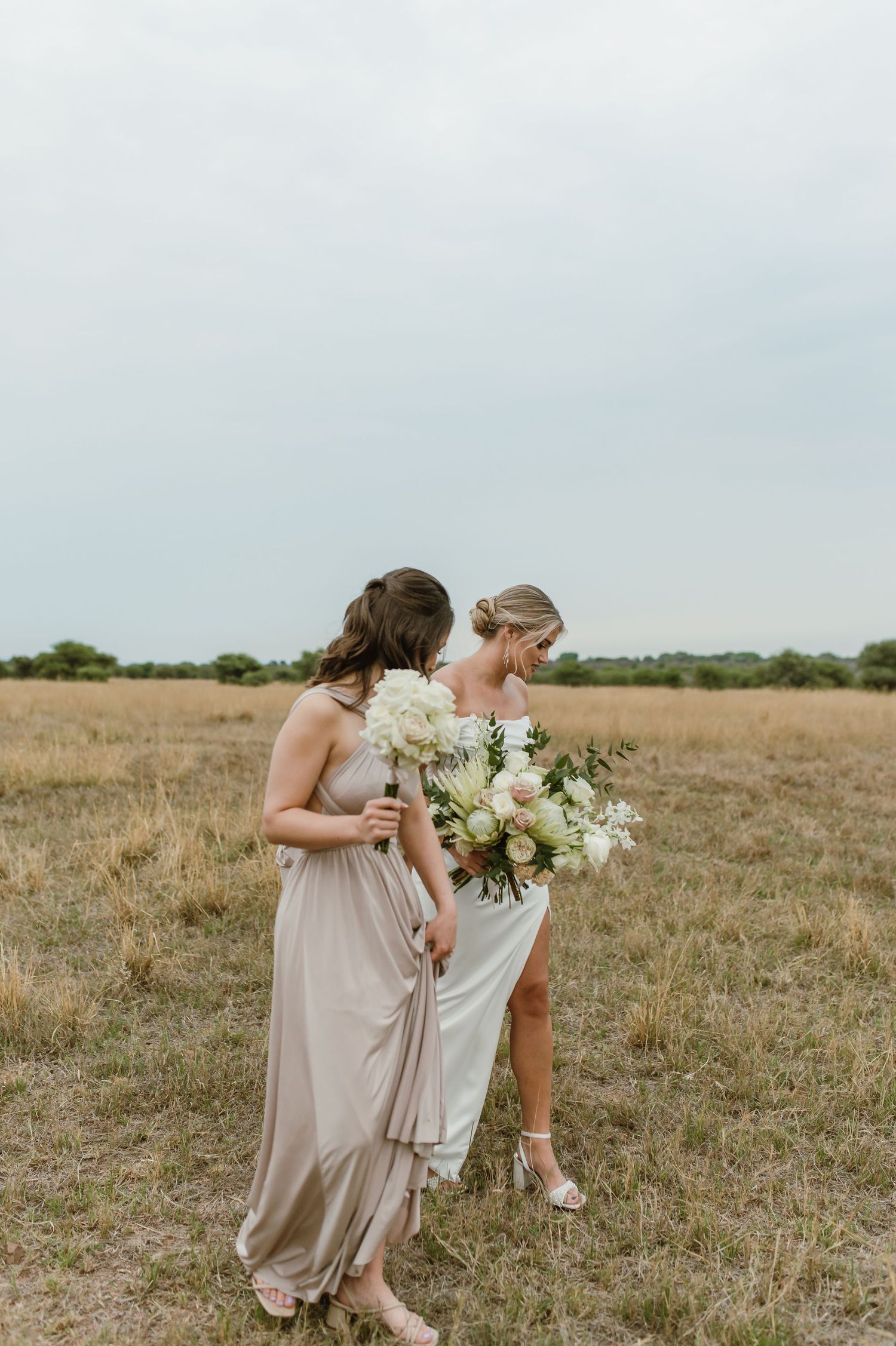 Bridesmaids in champagne dresses walk through a grassy field holding white floral bouquets.
