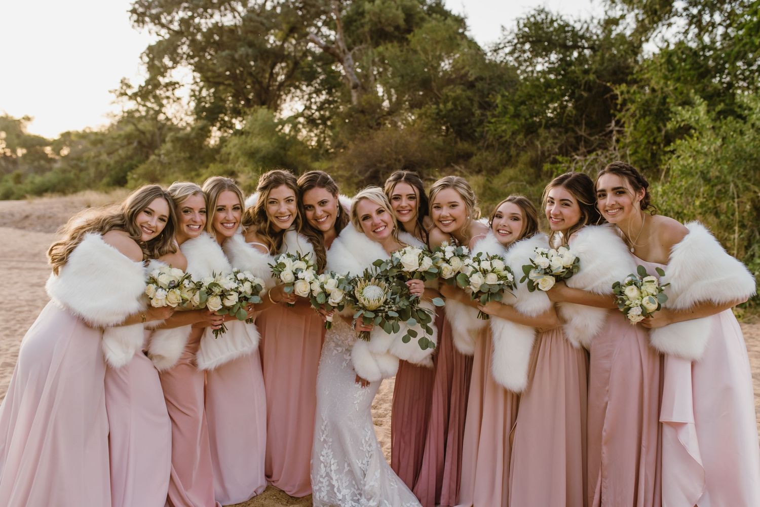 Bridesmaids in pink gowns pose together with floral bouquets on beach.