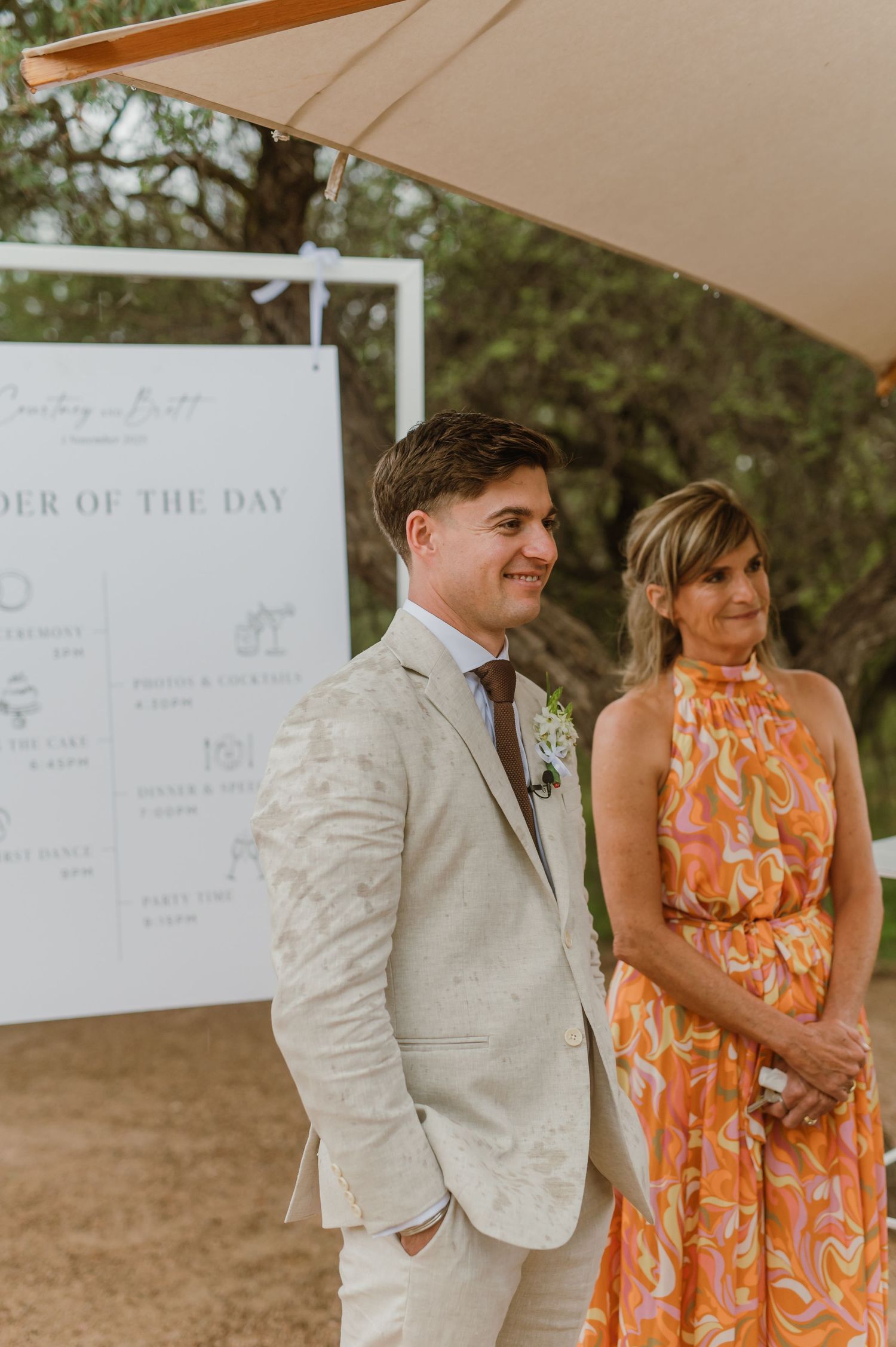 Wedding party members in light colored attire pose under a beige canopy with order of day signs visible.