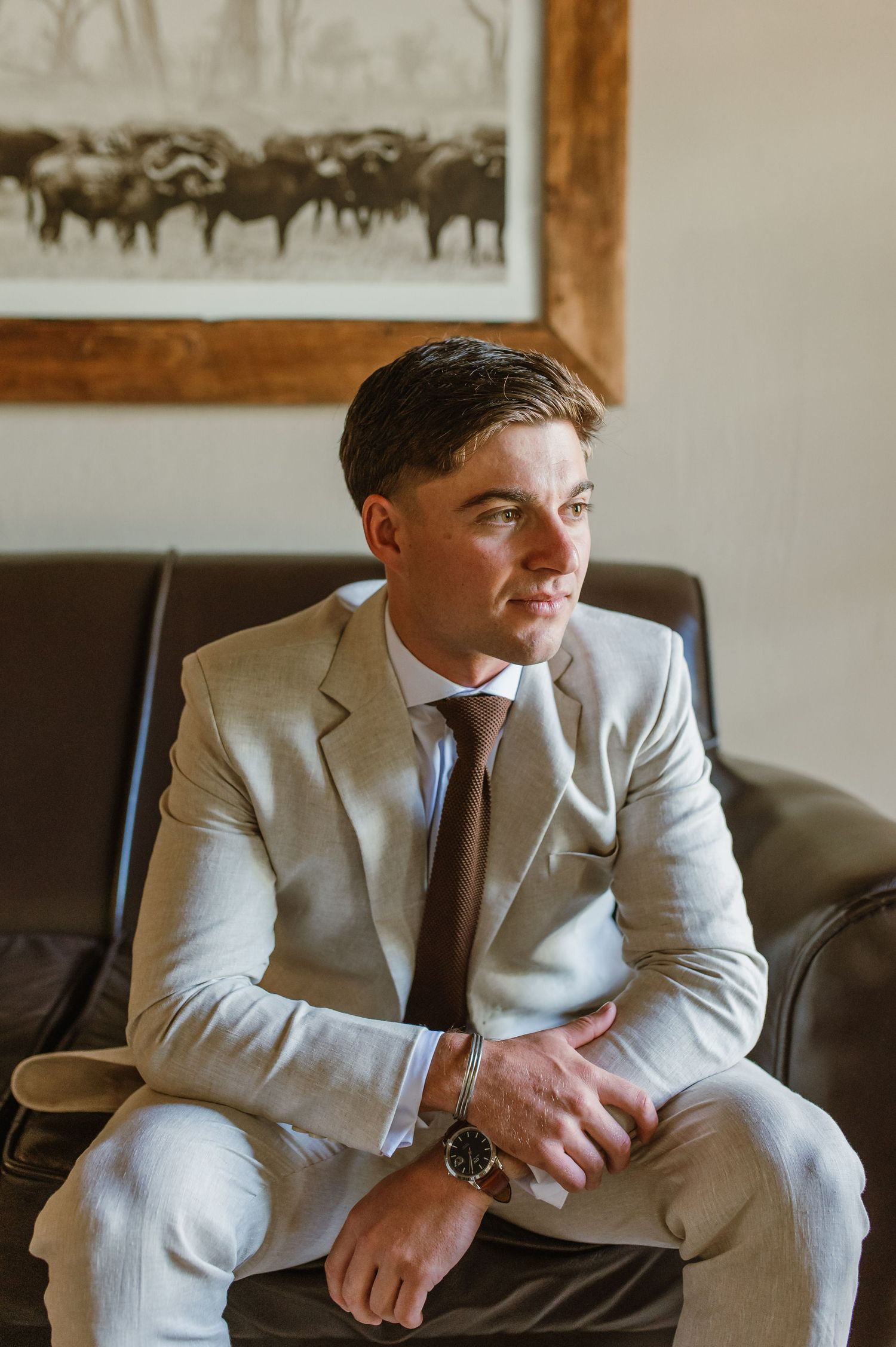 A person in a light grey suit sitting on a leather couch beneath framed artwork.