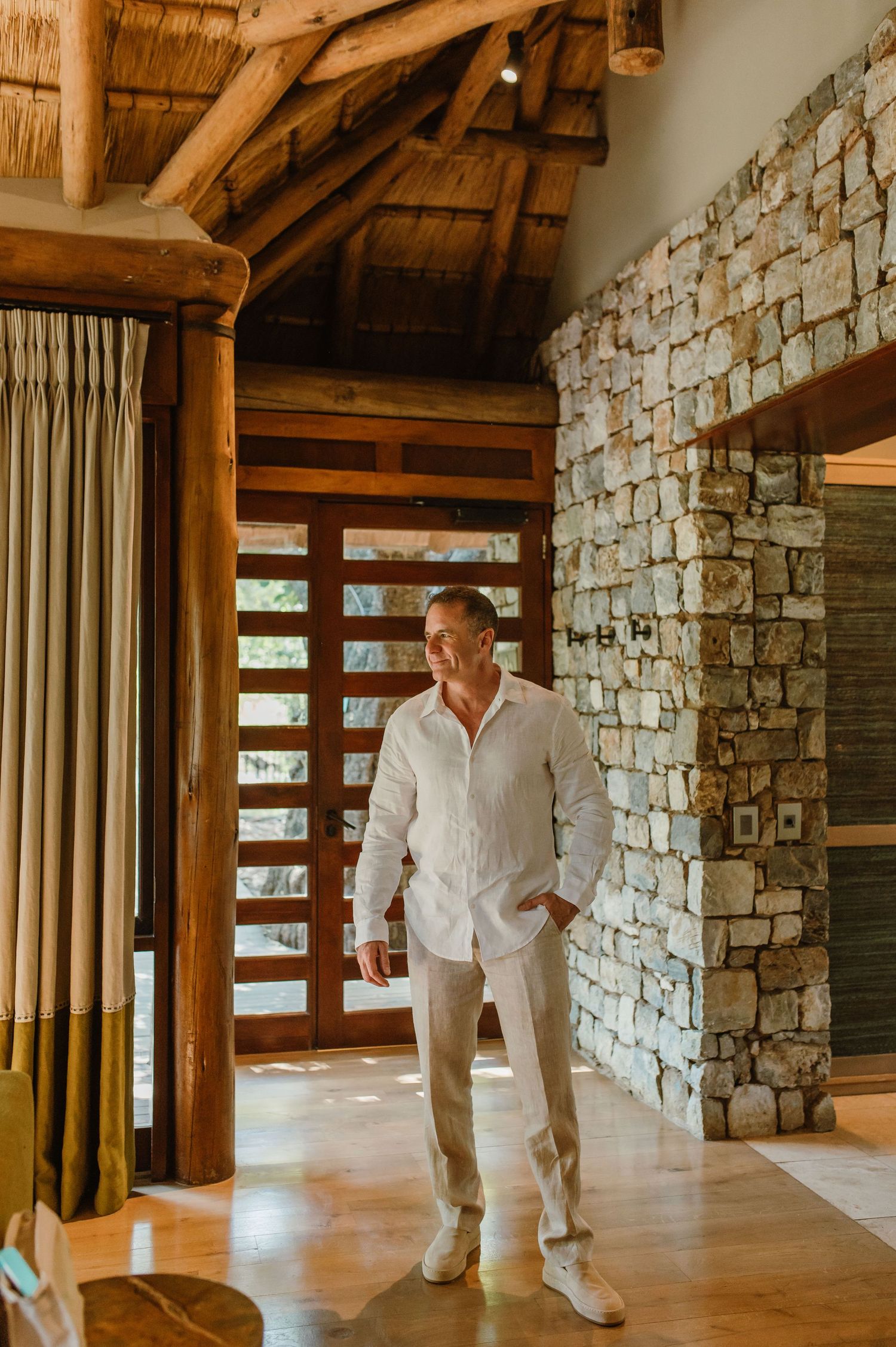Elegant man in white linen shirt standing in a luxury safari lodge with stone walls and thatched roof.