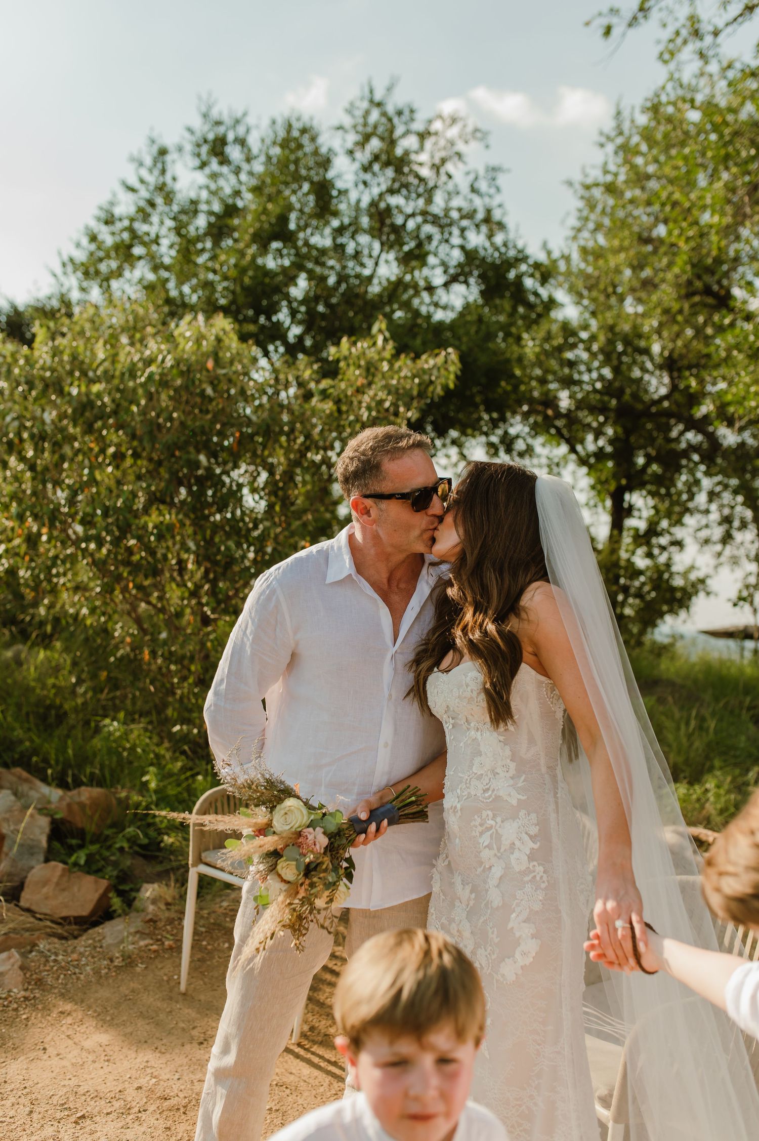 Newlywed couple kissing outdoors, bride in lace gown holding bouquet with child nearby.