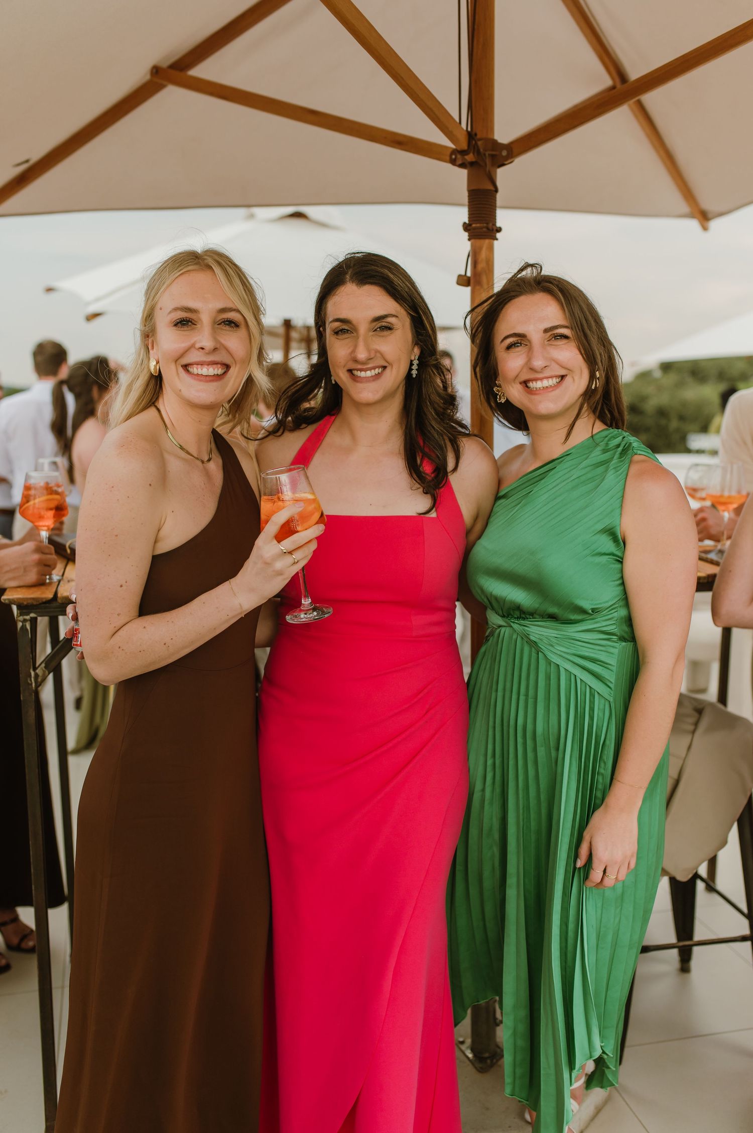 Three women in coordinating formal dresses in brown, pink and green smile together under white umbrellas at an outdoor event.