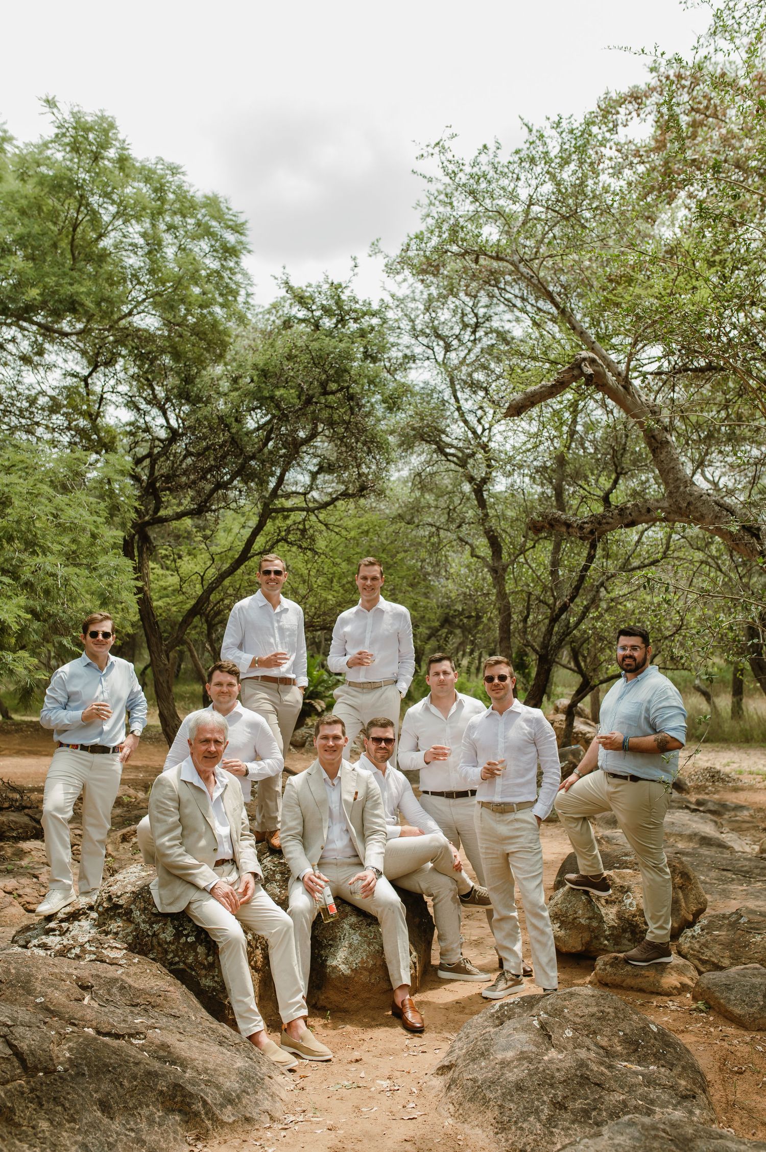 A group in light colored suits and shirts gather on rocky terrain under a canopy of trees for a casual photo.