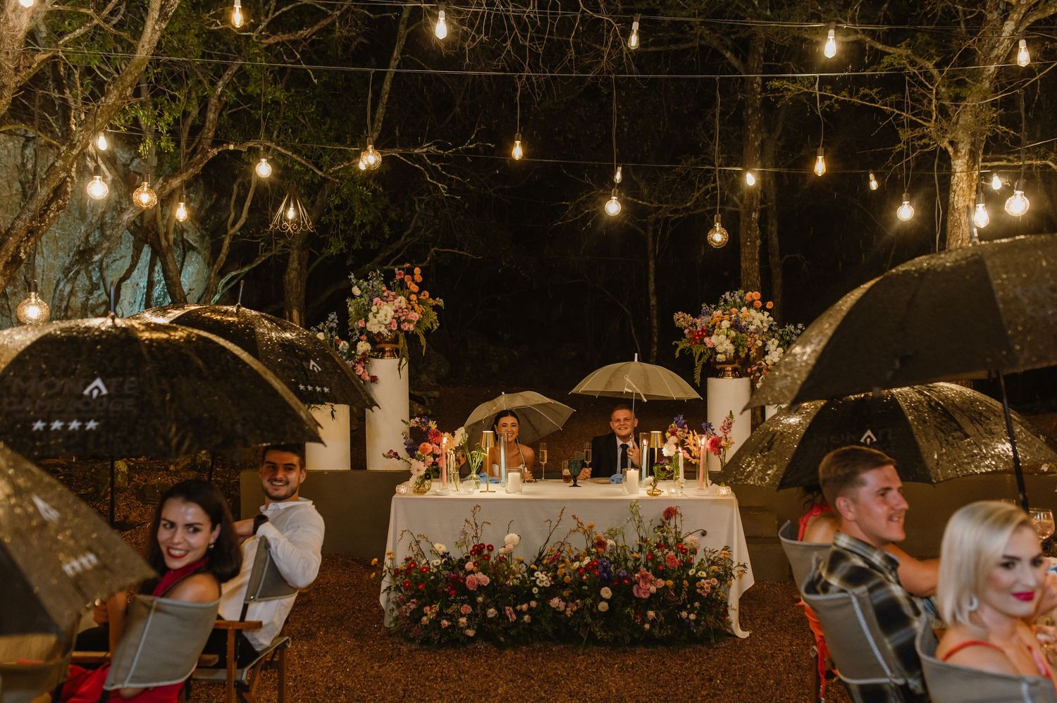 Wedding couple at sweetheart table under string lights as guests hold clear umbrellas in rain.