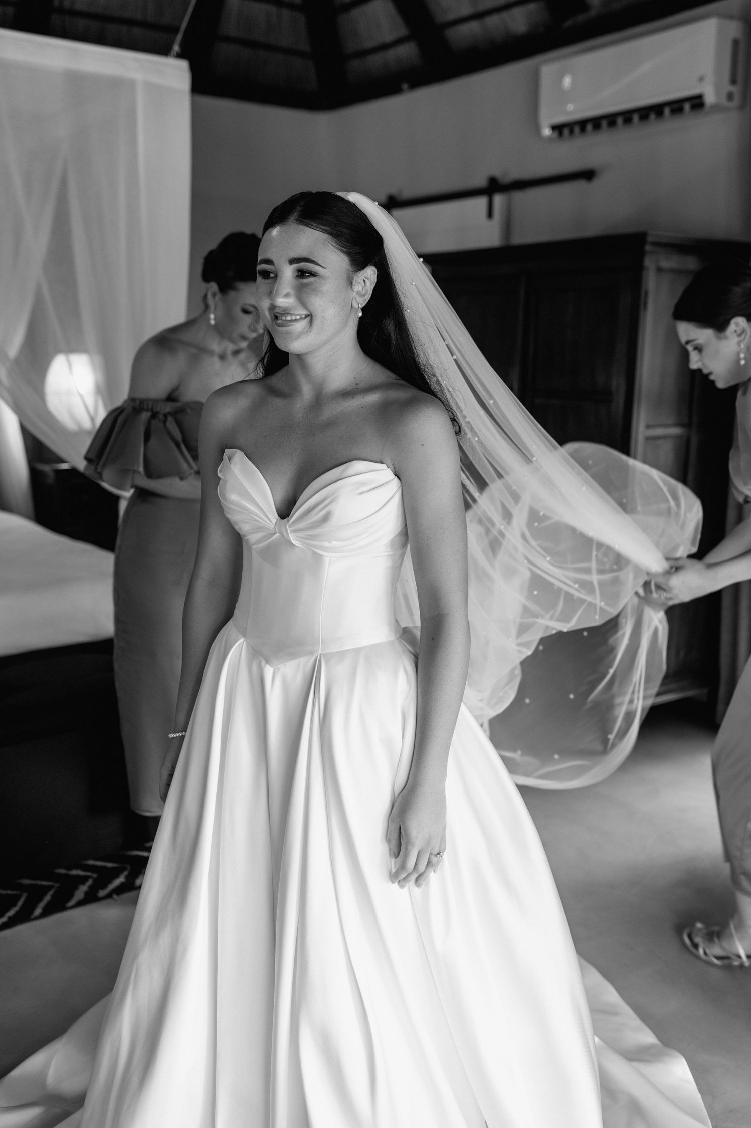 Black and white photo of smiling bride in strapless gown with veil being adjusted by bridesmaids.