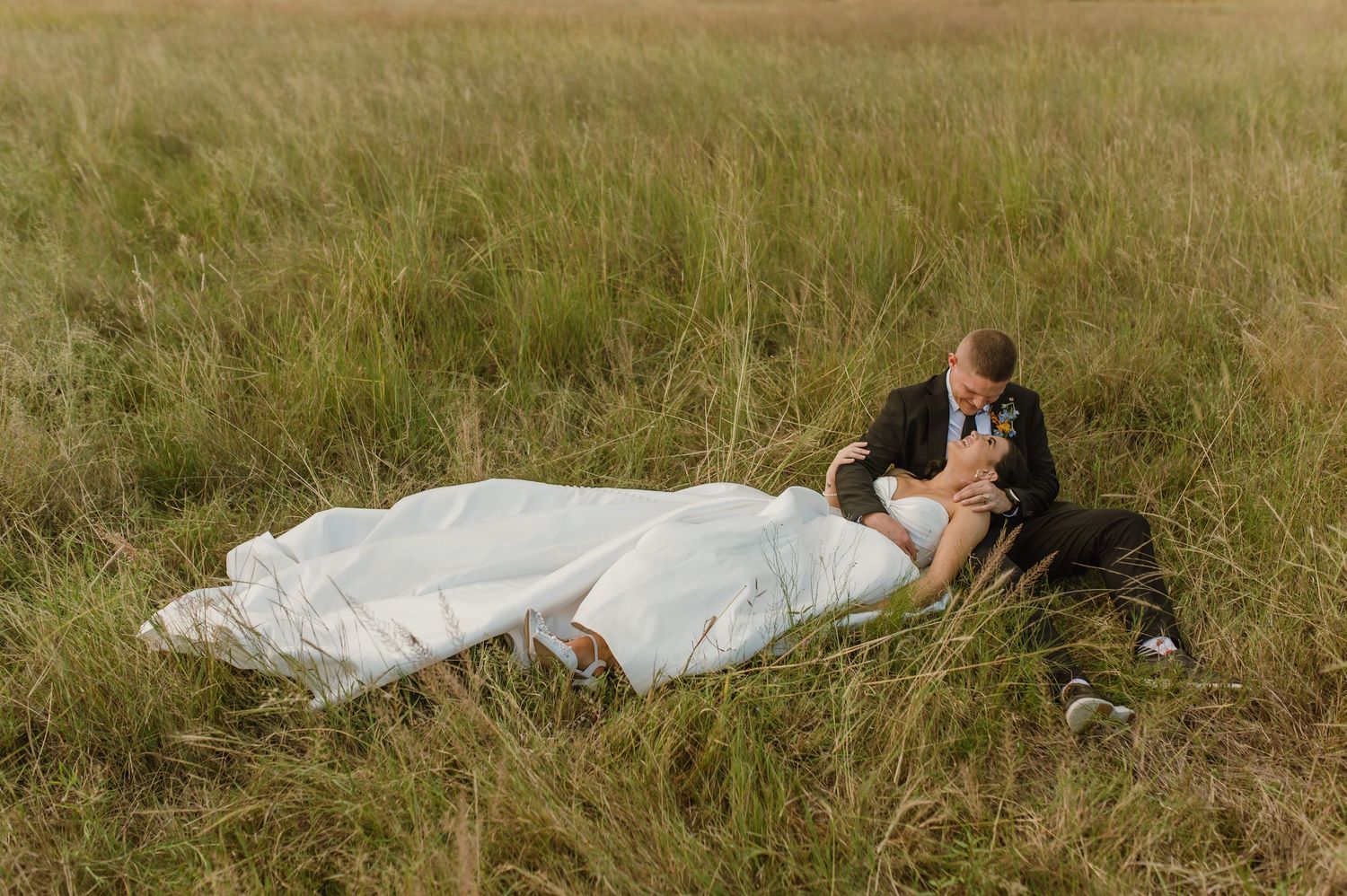 Bride and groom sharing a romantic moment lying together in a lush green grass field.