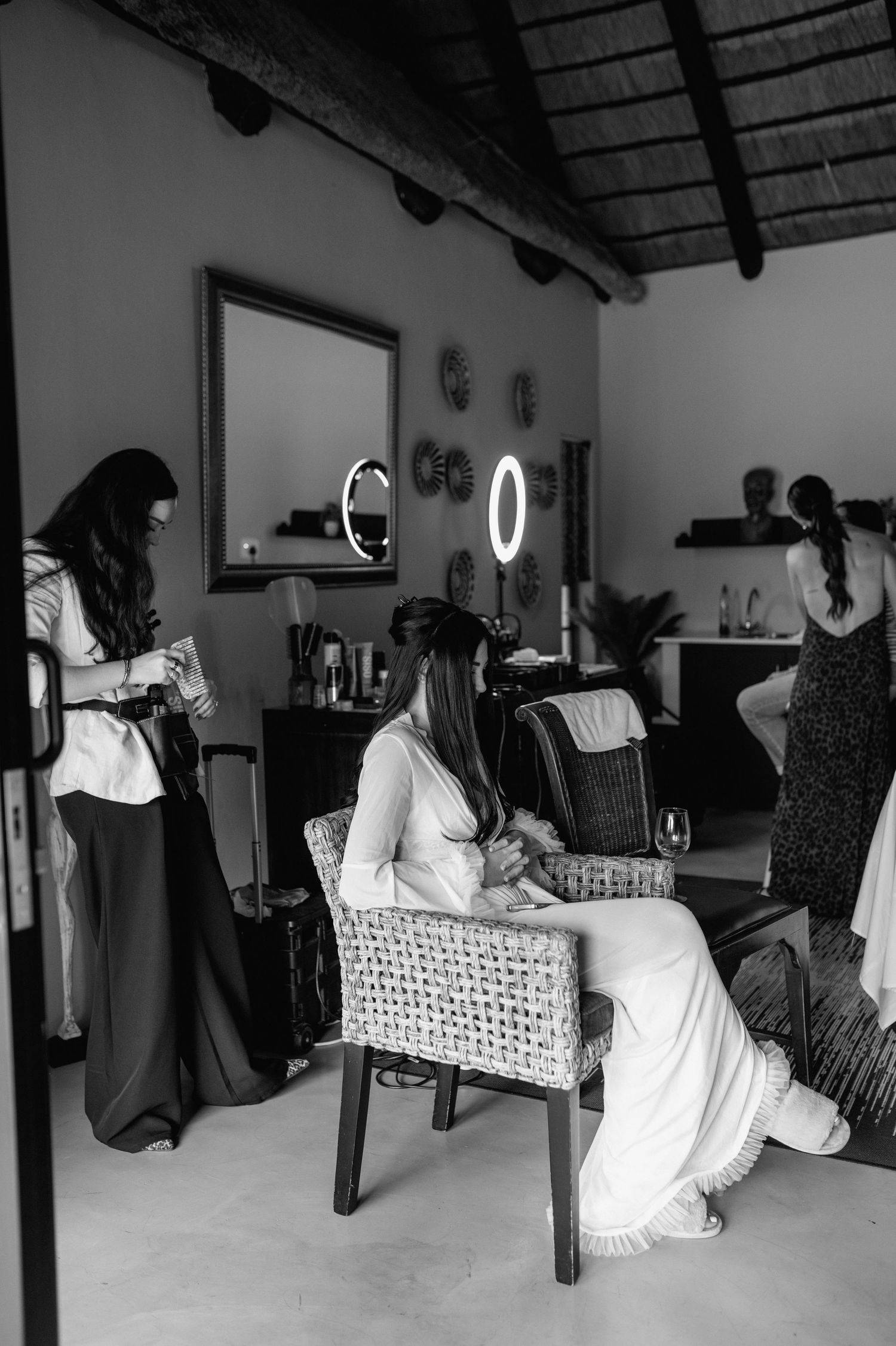 Bride getting ready in a rustic salon with a ring light mirror and attendants helping in black and white.