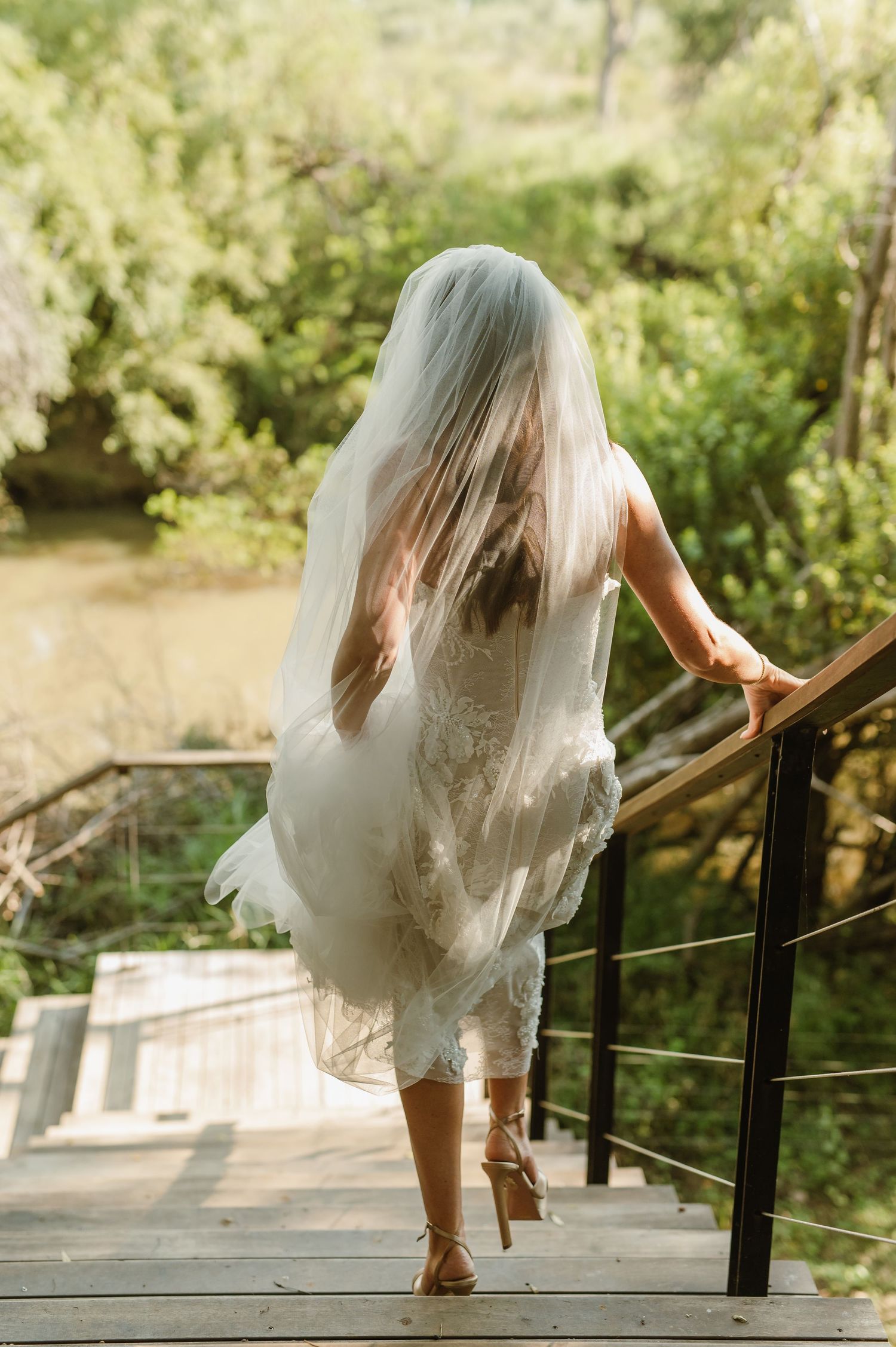 Bride in lace gown and veil walking up wooden stairs surrounded by greenery.