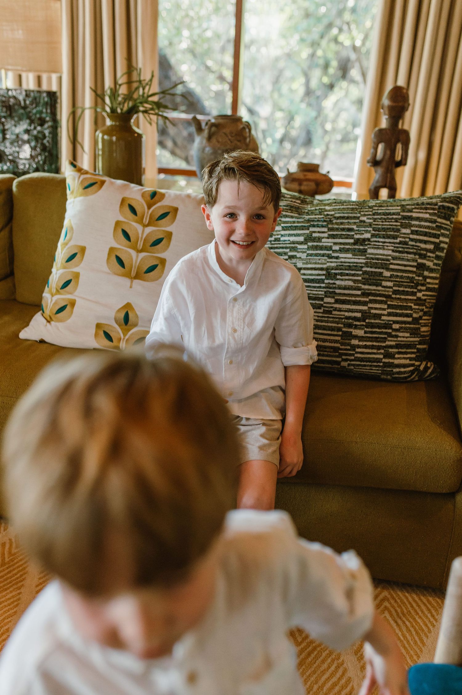 Smiling young boy in white shirt sitting on a green sofa in a warmly decorated safari lodge.