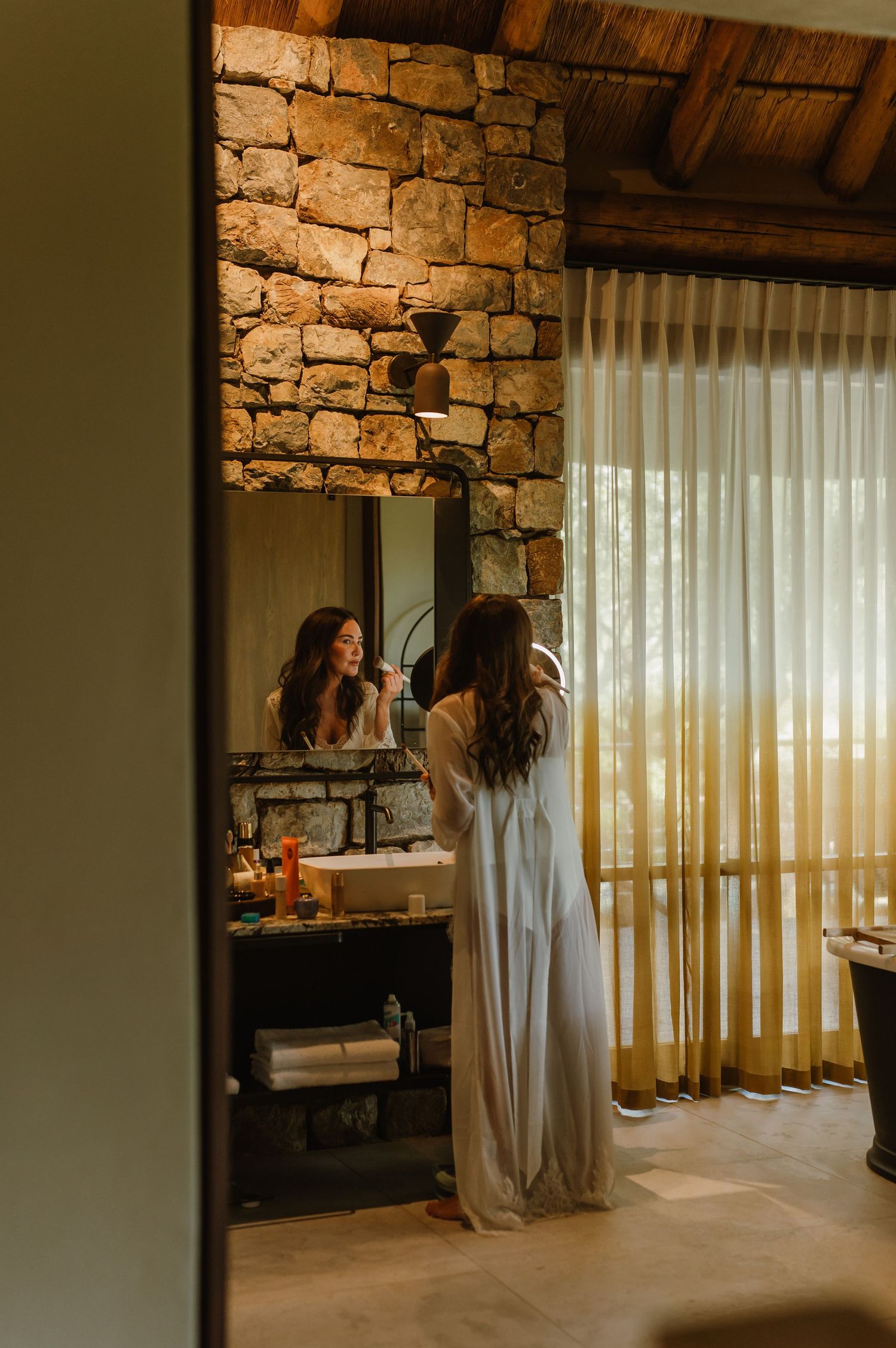 Bride in white robe doing makeup at a stone-walled bathroom mirror in a luxury safari lodge.