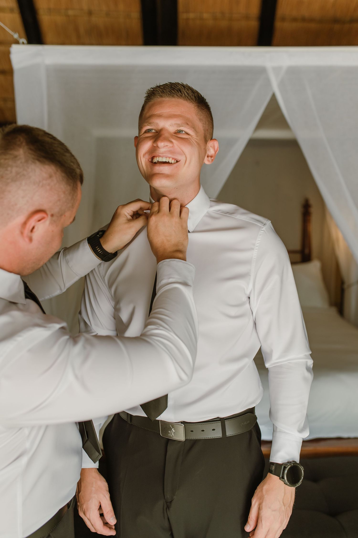 Groom laughing as his groomsman helps adjust his collar in a rustic thatched-roof lodge room before the wedding.