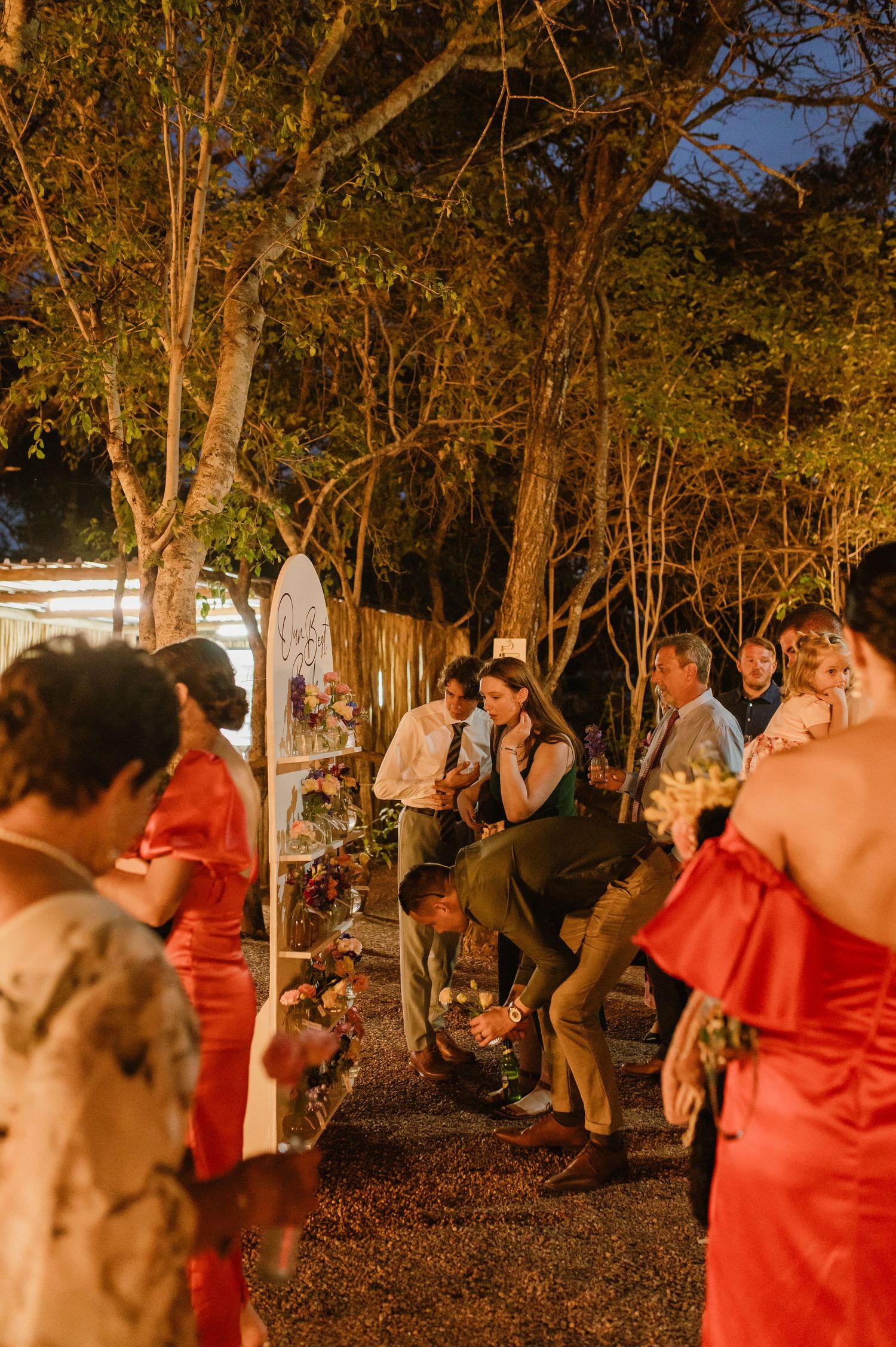 Wedding guests mingle and laugh at lively outdoor evening reception under illuminated trees.