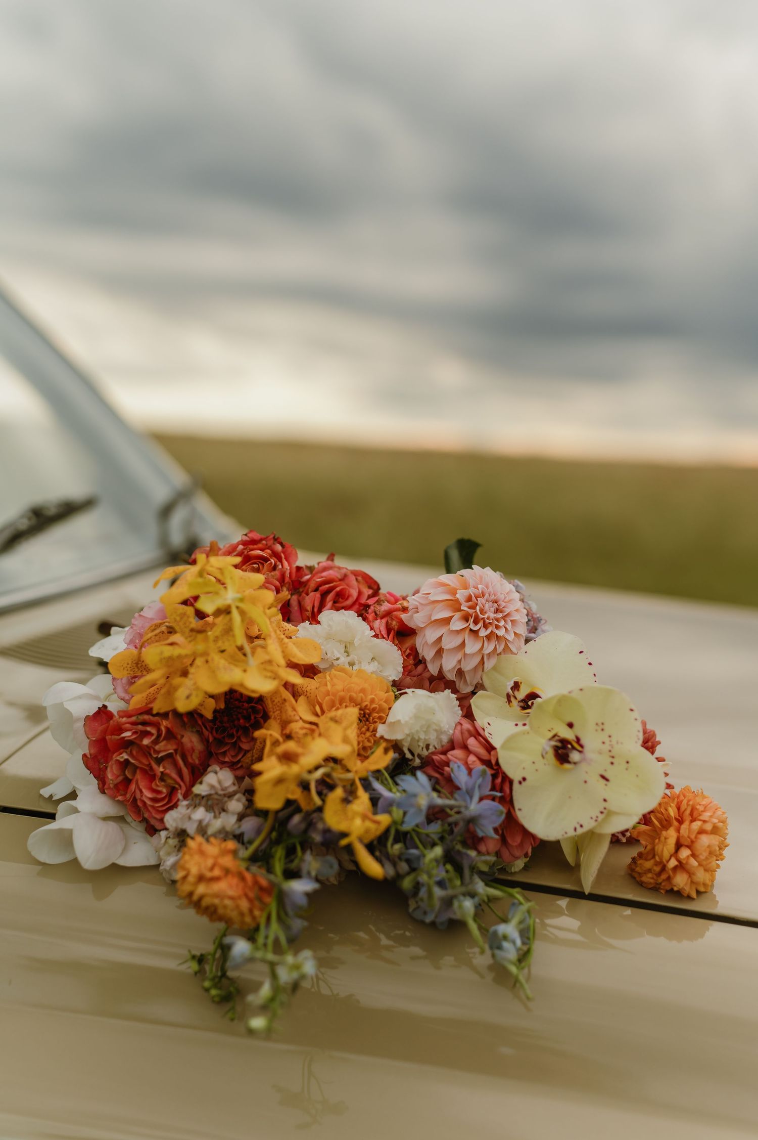 Colorful bridal bouquet with dahlias and wildflowers resting on vintage cream vehicle hood.
