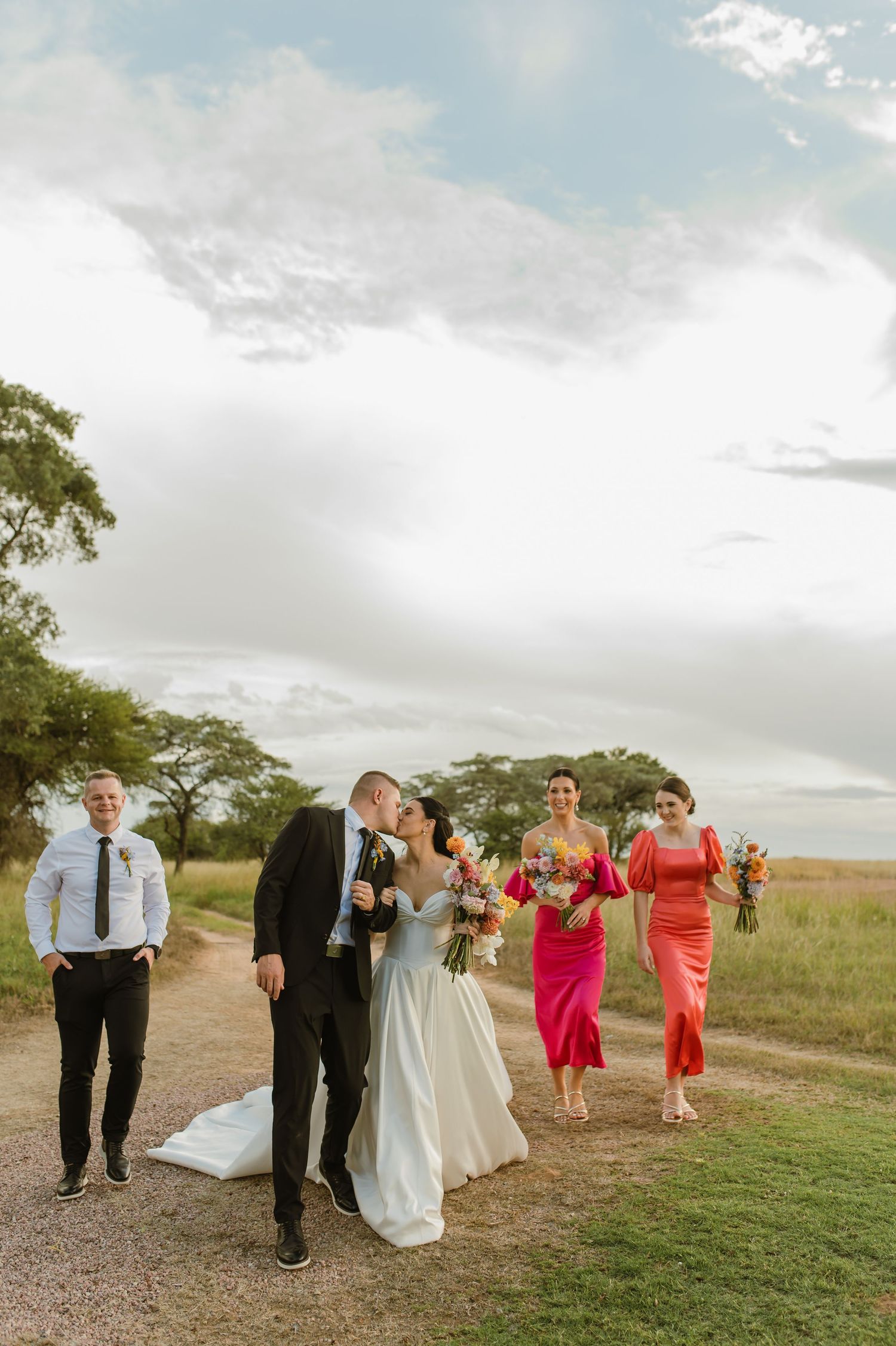 Bride and groom kiss while walking with bridal party in pink dresses along gravel path under dramatic sky.