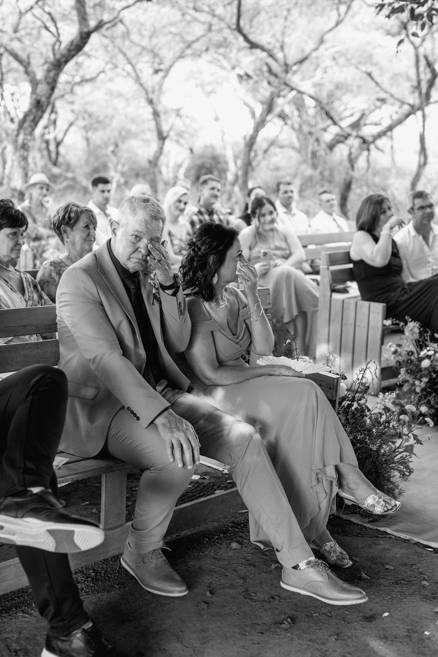 Black and white photo of wedding guests seated on benches sharing an intimate moment outdoors.