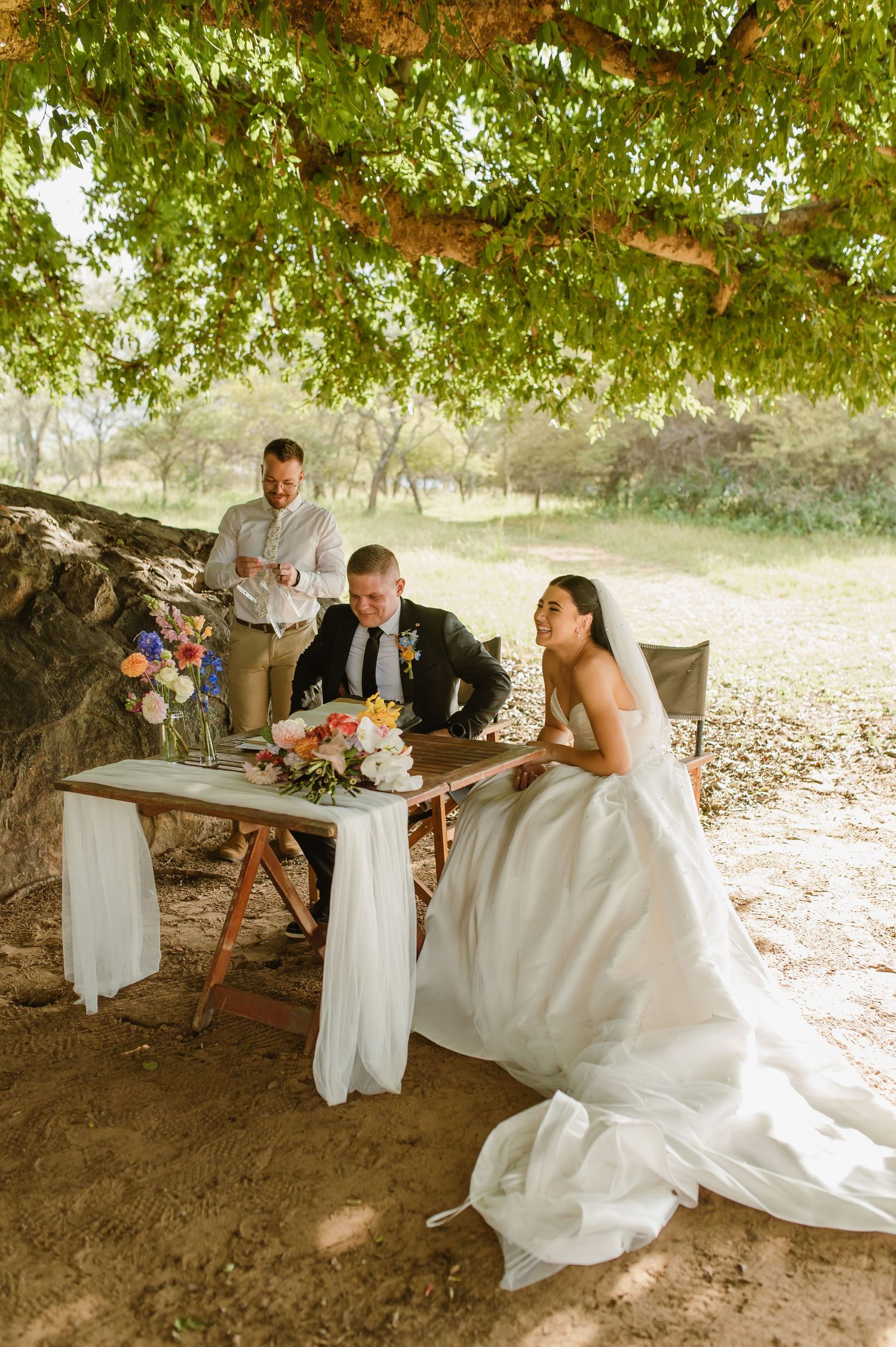 Bride and groom signing wedding documents at a rustic table under a large tree with an officiant standing nearby.