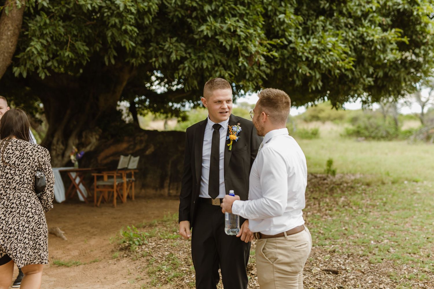 Two men in formal attire converse outdoors under a large tree at a wedding venue.
