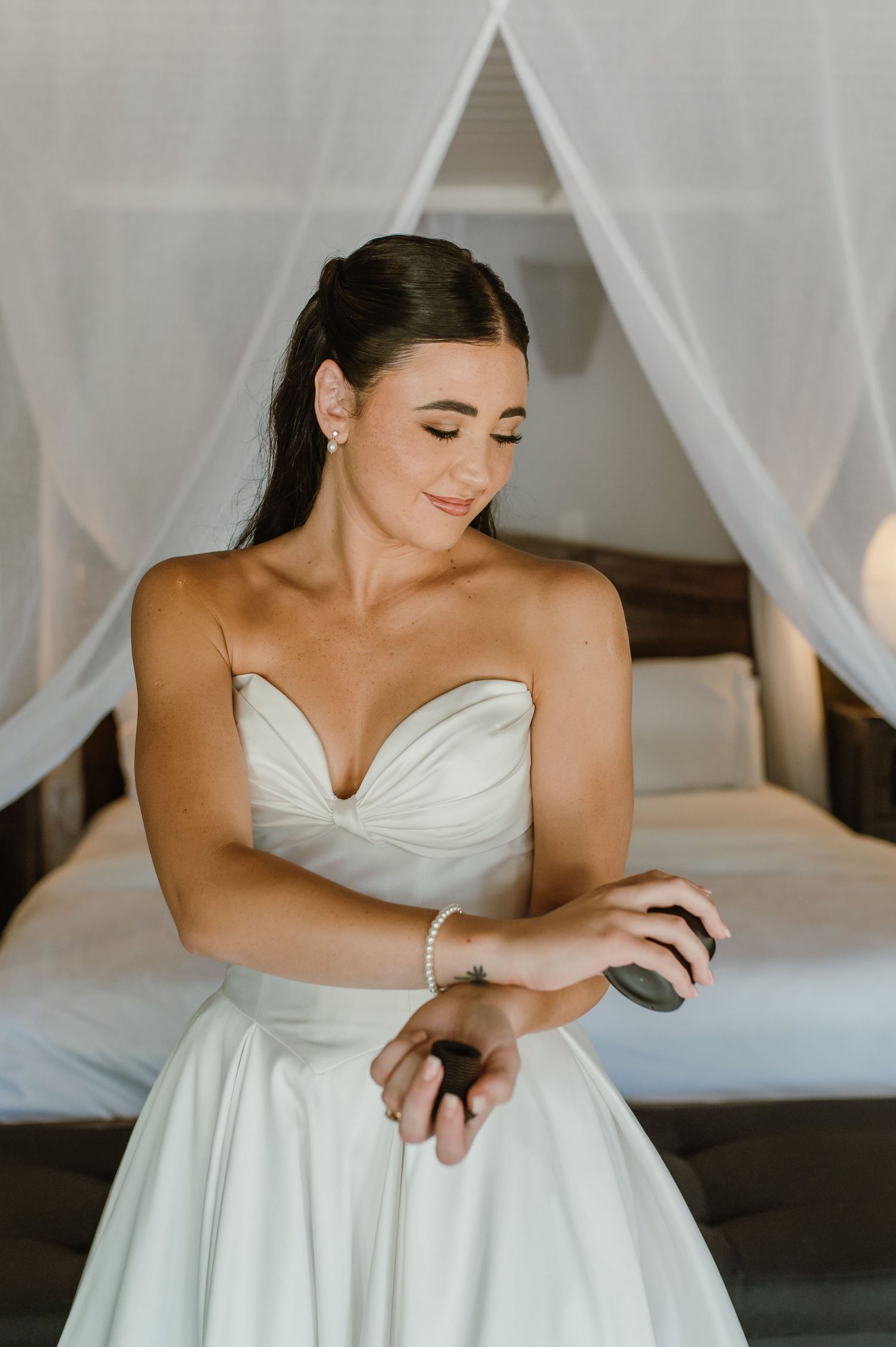 Bride in strapless white gown adjusting bracelet while seated near a canopy bed.