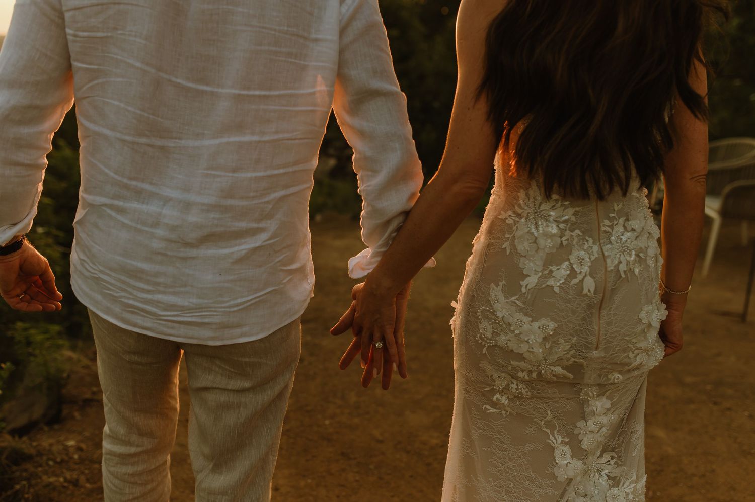 A bride and groom walk hand in hand in matching white and lace outfits at golden hour.